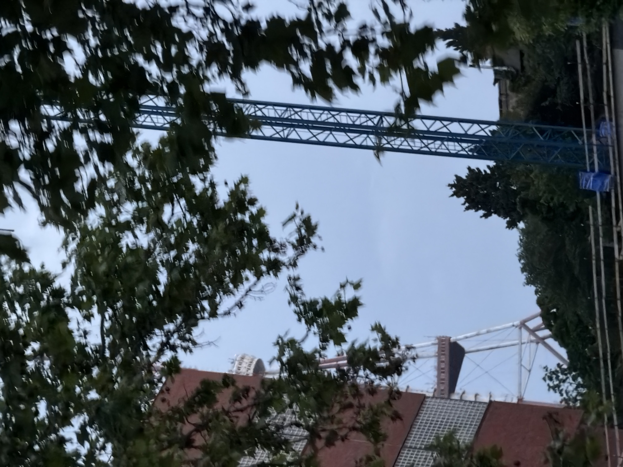 The image shows a partial view of the Bridge of Peace in Tbilisi, Georgia, as seen through trees. The bridge's steel structure is visible against a cloudy sky. The architecture below is a building with a red roof. Construction scaffolding and a crane are seen on the right of the image.