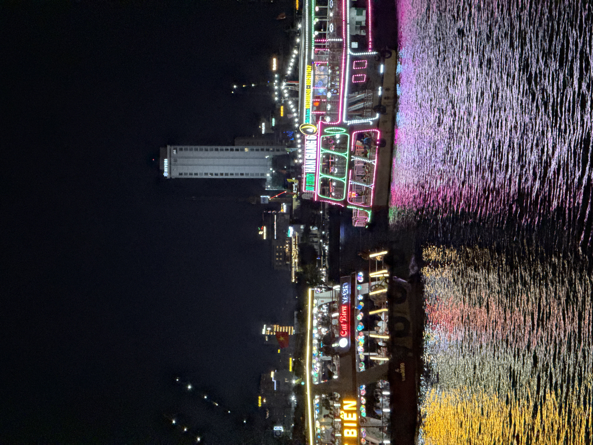 This media file depicts a vibrant night scene along the Hàn River in Đà Nẵng, Vietnam. The image is dominated by several large, brightly illuminated tour boats, docked or cruising on the dark water. These boats are extensively decorated with colorful neon lights, primarily in shades of pink, green, blue, and white, creating a festive atmosphere. Silhouettes of people enjoying the evening are visible on the decks of the boats, suggesting dinner cruises or evening entertainment.
The artificial lights from the boats cast striking, elongated reflections onto the river's surface, creating shimmering streaks of purple, pink, white, and golden yellow light that ripple across the water.
In the background, the city skyline is visible against the dark night sky. A tall, modern white building stands out, bearing the illuminated sign "HIYORI" near its top. Another building in the mid-ground has the word "CANVAS" displayed. Visible text on the boats includes "BIEN," "Cat Bien Gh" (likely Cát Biển), and "DU THIÊN HÀN GIANG 6" (meaning "Han River Cruise 6"), along with a phone number "0935888246-09". The overall impression is one of a lively, bustling tourist destination at night, showcasing the city's illuminated waterfront.