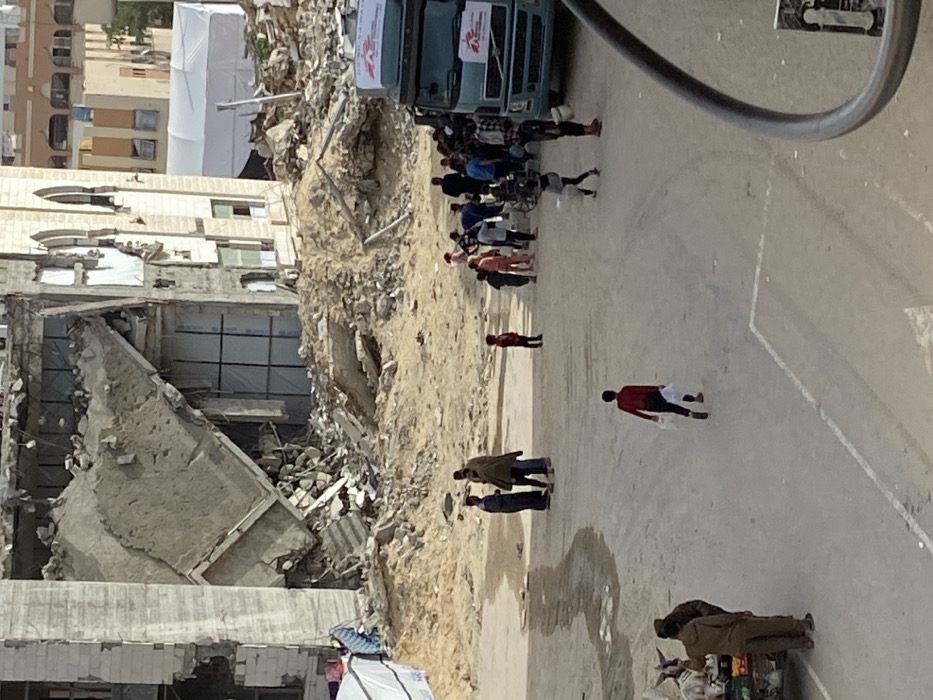 The image depicts a devastated urban landscape in Khan Yunis, Palestinian Territory. In the background, severely damaged multi-story concrete buildings stand partially collapsed, with exposed rebar and rubble. One structure exhibits distinctive arched window frames on its upper floors, adjacent to a section with significant structural failure and debris. Large mounds of dirt and rubble are piled around these ruined buildings. A white tent structure is visible beyond the damaged buildings in the far background.
In the midground, an unpaved, sandy-dirt area with some wet patches dominates the space between the rubble and the foreground. A light blue/green truck, prominently displaying "MEDECINS SANS FRONTIERES" (Doctors Without Borders) and its logo, is positioned on the right side of this dirt area, facing left. A crowd of approximately 15-20 individuals, comprising adults and children, are gathered around the truck, appearing to collect or receive items; several individuals are seen holding white buckets and plastic containers. To the left of this gathering, three adults are standing together, engaged in apparent conversation. A lone child in a red top stands further left in the dirt area.
In the foreground, a paved or hard-packed dirt surface shows vehicle tracks. A child in a red top and dark pants walks away from the center, carrying two white plastic bags in their left hand. On the far left, two adults are positioned near a small, multi-tiered cart. A dark metal railing is visible on the right edge of the frame.