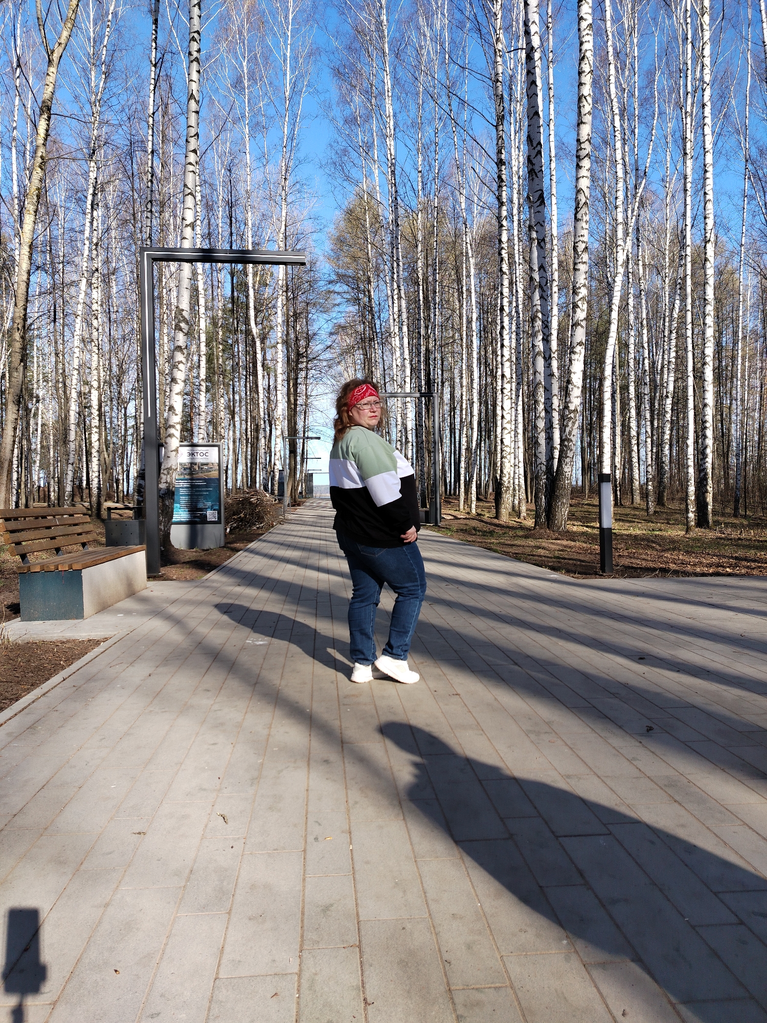 A person stands on a paved path in a birch tree forest. The path is made of gray rectangular pavers. The person has light skin and is wearing a red patterned bandana, a black, white, and green sweater, blue jeans, and white sneakers. The person is looking over their shoulder towards the left of the frame. To the left of the person is a metal sign with text and images on it, next to a wooden bench. Tall birch trees with bare branches fill the background and sides of the frame. The sky is blue and visible through the trees. The light suggests it is daytime. Long shadows are cast on the path. The image was taken in the city of Chaikovsky, Russia.