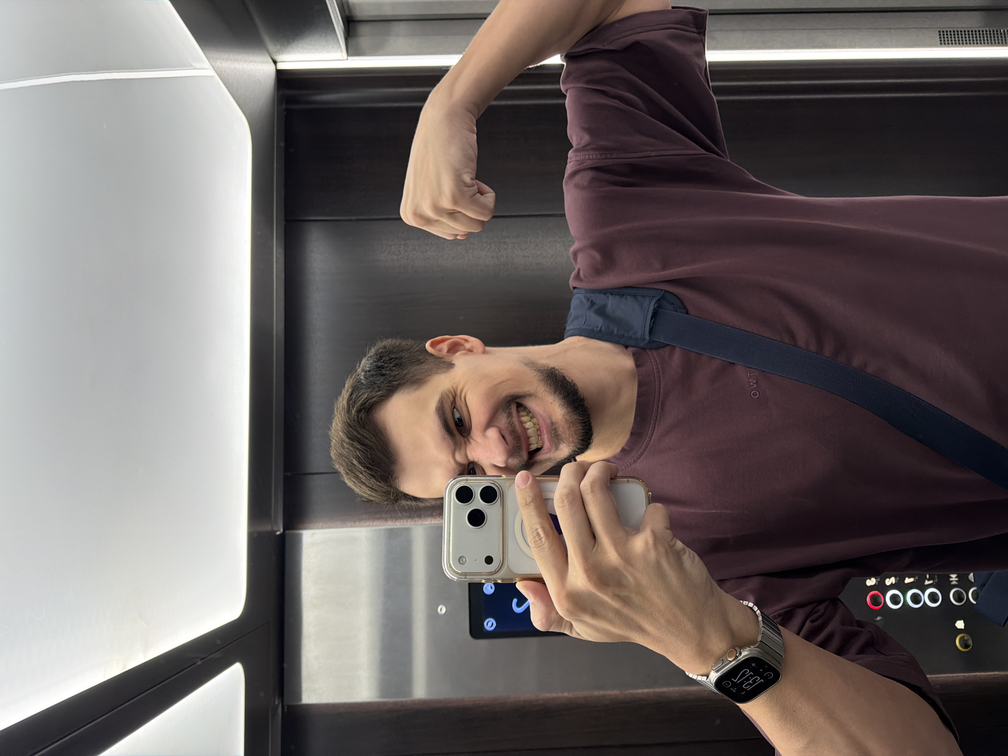 A man is taking a selfie in an elevator in Krasnoyarsk, Russia. He is wearing a burgundy t-shirt, and a silver Apple Watch. The elevator has stainless steel walls and black accents. The buttons of the elevator panel are visible. A white panel with an integrated light is on the left wall. The man is flexing his bicep in the photo. He is wearing a navy blue bag strap across his chest.