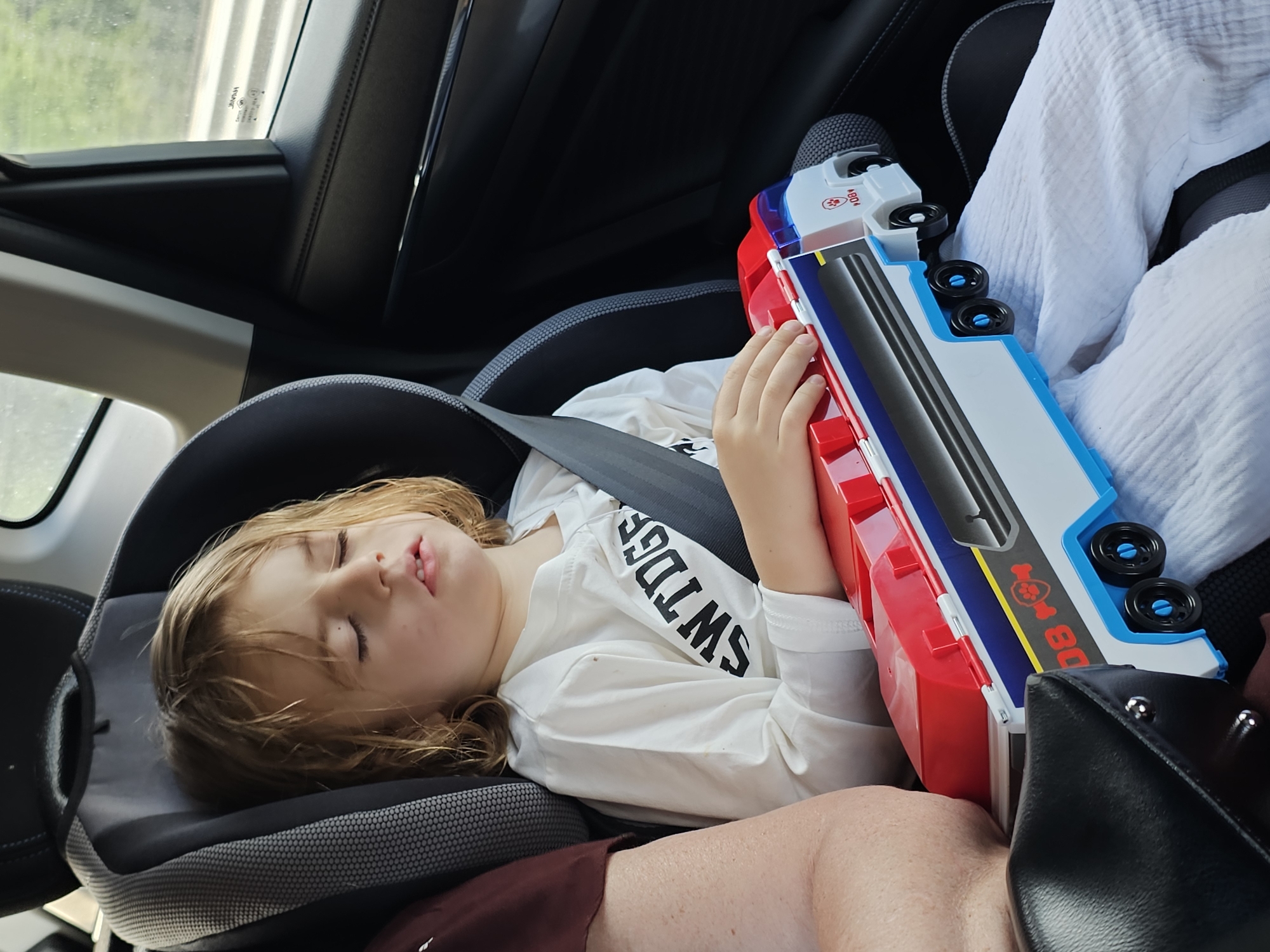 A young child with blond hair is sleeping in a car seat, restrained by a seatbelt. The child is wearing a white long-sleeved shirt. The child is holding a toy truck, primarily red, white, and blue. The car's interior is visible, including the headrest of the seat and the window. The location and city of Stimul, Russia, cannot be determined from the image.