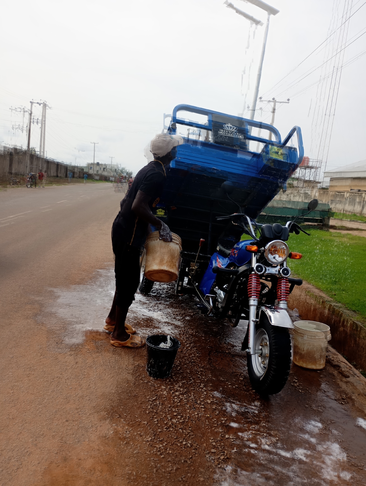 In Shendam, Nigeria, a dark-skinned person wearing a black t-shirt, black trousers, and light-brown sandals is standing on a wet, muddy road, holding a light-brown bucket filled with water. They appear to be washing a blue three-wheeled motorcycle with a raised cargo bed, which is parked next to a muddy embankment and a patch of green grass. The motorcycle has a silver fender, red suspension forks, and a chrome headlight. A black bucket sits on the ground near the person's feet, and a partially submerged bucket of water is visible on the embankment. The sky is overcast, and there are power lines and utility poles in the background. A concrete wall runs along the side of the road, with some buildings visible in the distance. The overall scene suggests an outdoor washing activity in a somewhat rural or developing urban environment.