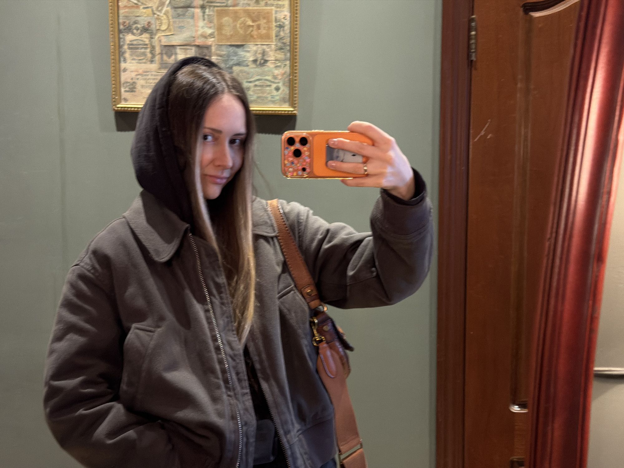 A young woman with light brown hair and brown eyes is taking a selfie in what appears to be a room with drab green walls. She is wearing a charcoal gray jacket with a hood pulled up, a brown leather purse with a wide strap is hanging from her shoulder. The woman is holding an orange iPhone with a multi-colored patterned case. Behind her is a framed picture on the wall and a wooden door with a red curtain. There is no information given to suggest that the location is in the city of Khimki, Russia.