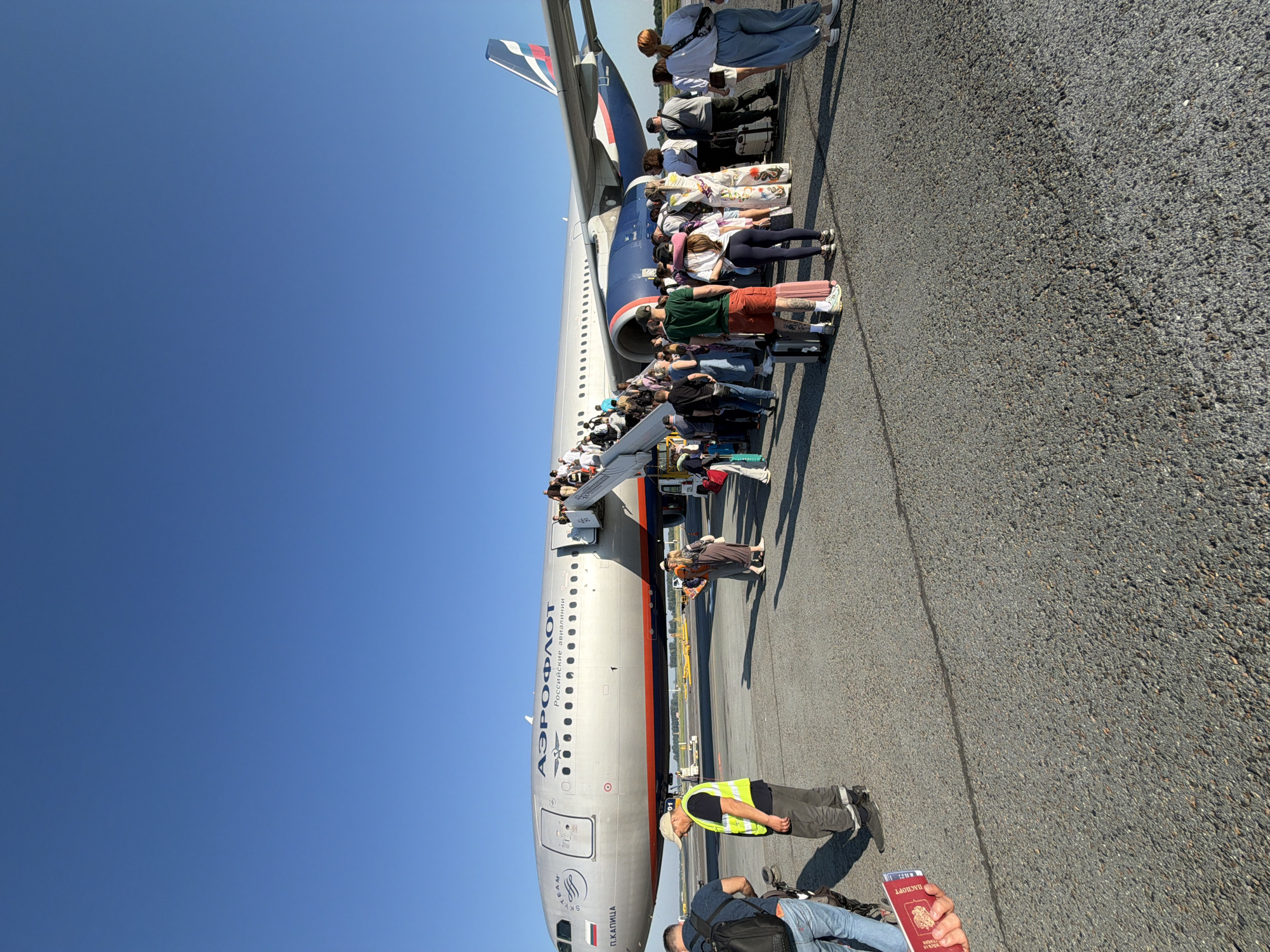 An Aeroflot airplane is parked on an asphalt tarmac under a clear blue sky. Passengers are disembarking the aircraft via a mobile stairway. Several individuals are on the ground near the plane, some carrying luggage. One person in the foreground is holding a red passport. The scene appears to be at an airport in the city of Ob, Russia, as suggested by the Aeroflot branding on the aircraft and the context of the location.