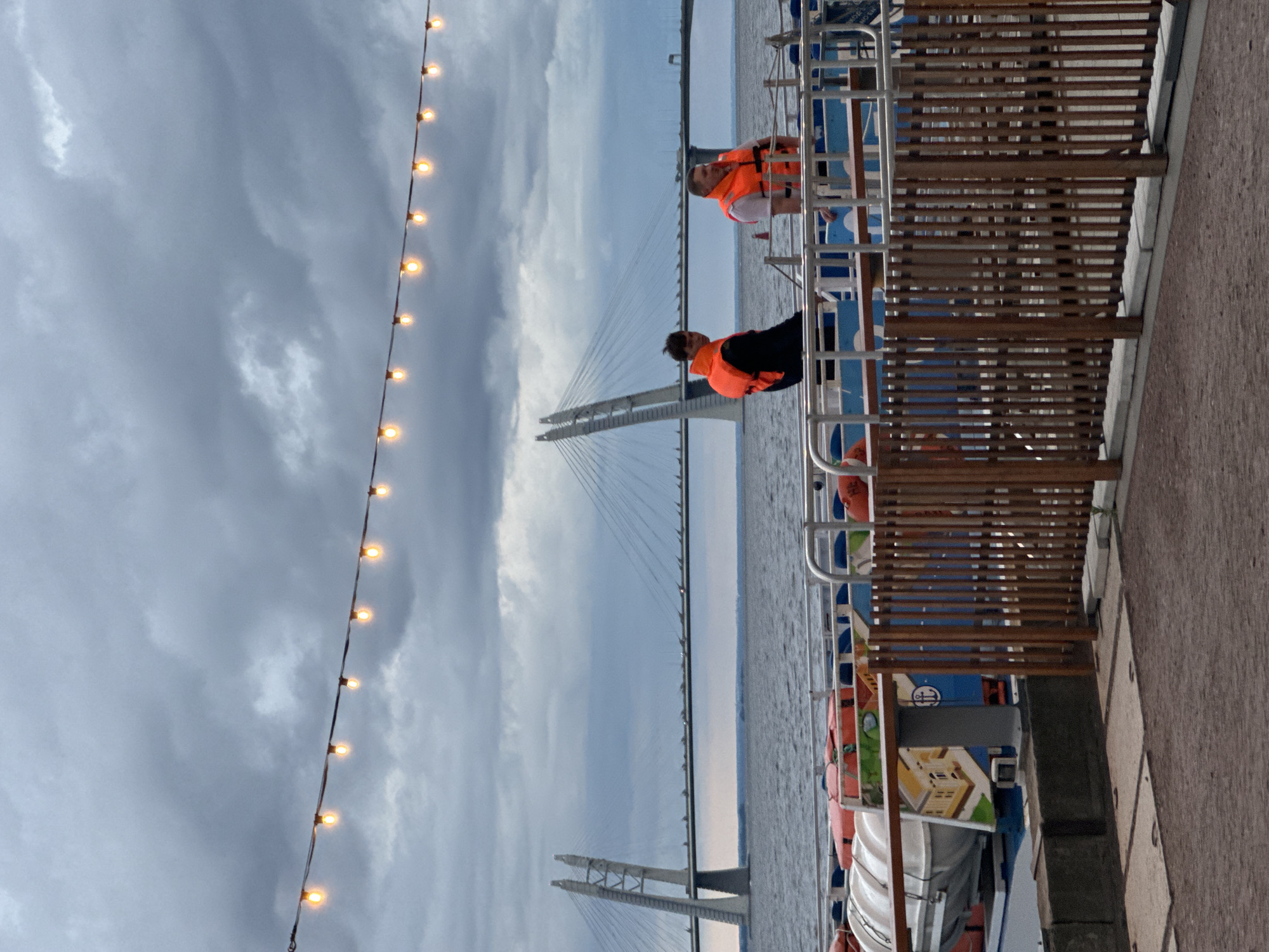 The image shows a cloudy day with a cable-stayed bridge in the background. In the foreground, two men wearing orange life vests stand on a boat deck. The boat is docked by a promenade with a wooden fence. The bridge is likely the Western High-Speed Diameter bridge in Saint Petersburg, Russia, known for its distinctive cable design and its role in connecting different parts of the city across the Neva Bay. The lighting suggests it's either early morning or late evening, or a particularly overcast day.