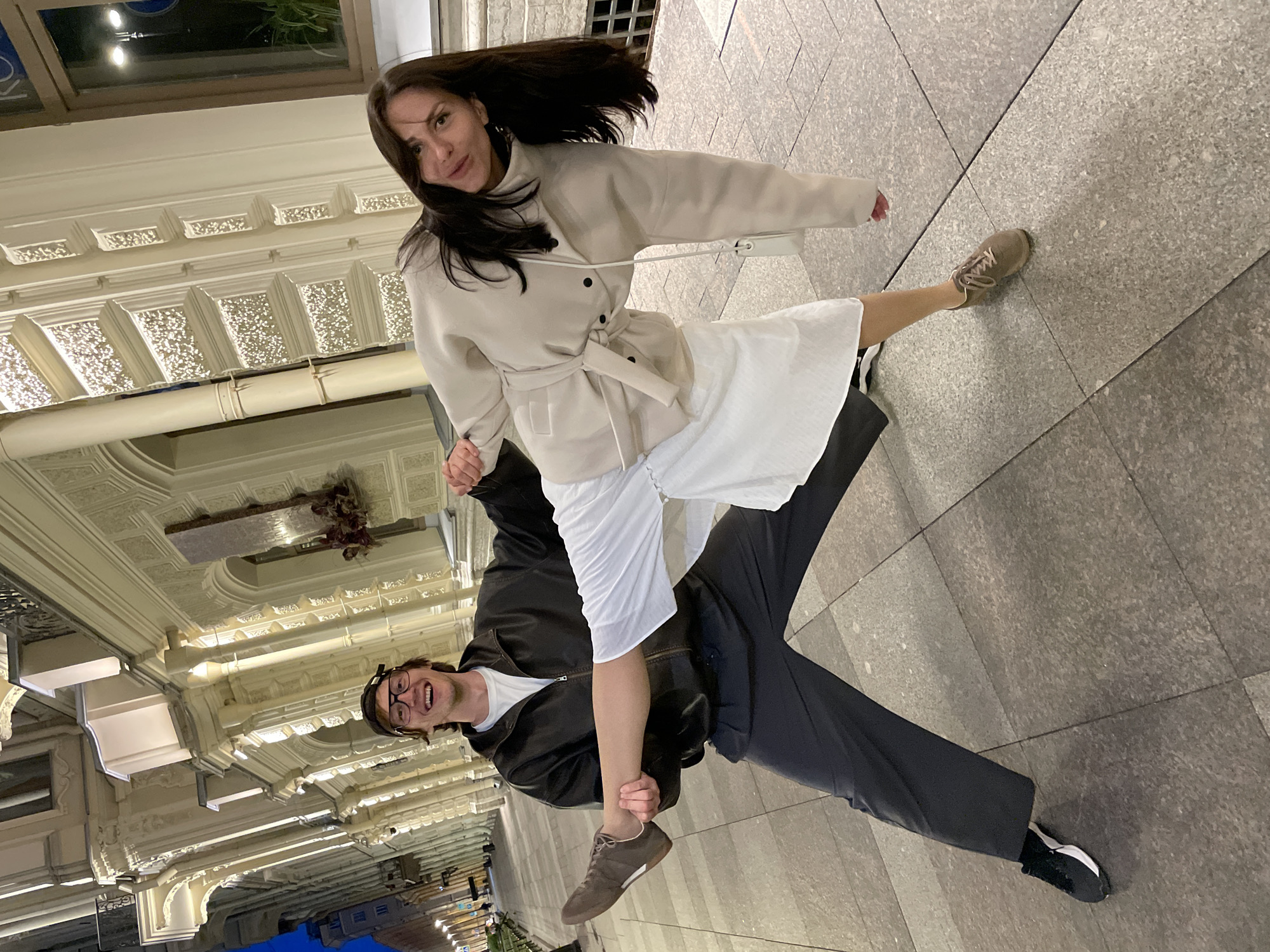 A man and a woman are posing for a photo in front of a building in Saint Petersburg, Russia, at dusk. The man, wearing glasses, a black leather jacket over a white t-shirt, and dark pants, is holding the woman's raised leg. The woman, with long dark hair, is wearing a light-colored trench coat over a white dress. She is leaning forward and looking at the camera. The building behind them is illuminated and features ornate architectural details, including columns and decorative carvings. The ground is paved with large, square tiles. The scene suggests a playful or artistic interaction between the two individuals.
