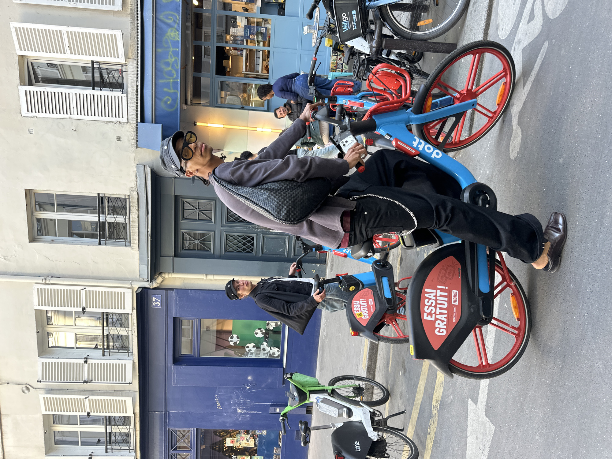 This image captures a vibrant street scene in Paris, France, featuring several people and rental bicycles amidst historic European architecture. The perspective is from slightly below, looking up at a person seated on a bicycle.
The main subject, a person wearing dark sunglasses, a dark cap, a grey long-sleeved shirt, and black pants, is seated on a bright blue and red Dott rental bike. They hold the handlebars, looking upwards and slightly to the left. A black woven bag is slung across their chest. Another person is visible behind them, also on a blue and red bike, wearing a dark jacket. Further in the background, more individuals are seen near other bicycles and a storefront.
The street features a grey pavement with yellow double lines and white markings that indicate a bicycle lane. Various rental bicycles are parked or in use, including blue and red Dott bikes, a green and white Lime bike, and a darker Véligo bike in the background.
The setting includes two distinct buildings. To the left, a light-colored building with multiple windows featuring white louvered shutters and wrought-iron window guards. The number "37" is visible on this building. Adjacent to it on the right is a dark blue building with a large storefront window and a blue awning, also showing graffiti that reads "RACUL!!". The time appears to be daytime, with clear weather and ample light.
Visible text includes brand names like "dott," "Véligo France," and "Lime" on the respective bicycles. Promotional text on the red wheel guards of the Dott bikes states "ESSAI GRATUIT!" (Free Trial!) and "ENTREZ LE CODE HIBIKE" (Enter the code HIBIKE), along with "HIBIKE." The building number "37" and the graffiti "RACUL!!" are also clearly visible.