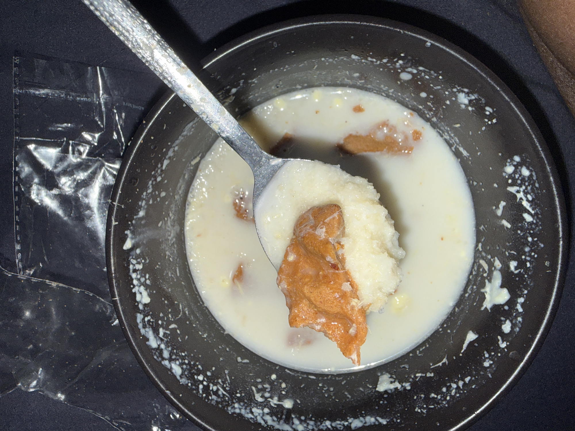 A close-up overhead shot captures a dark bowl of a milky dessert containing chunks of fried dough. A silver spoon is in the process of lifting a piece of the dough, revealing the creamy texture of the dessert beneath. The bowl's rim is speckled with white drops of the dessert, indicating it has been partially consumed. The background is a dark, textured surface, possibly fabric, with a crinkled clear plastic bag partially visible in the upper portion of the frame. There is no indication of the specific location within Abuja, Nigeria, or any discernible environmental features that would suggest a particular setting within the city.