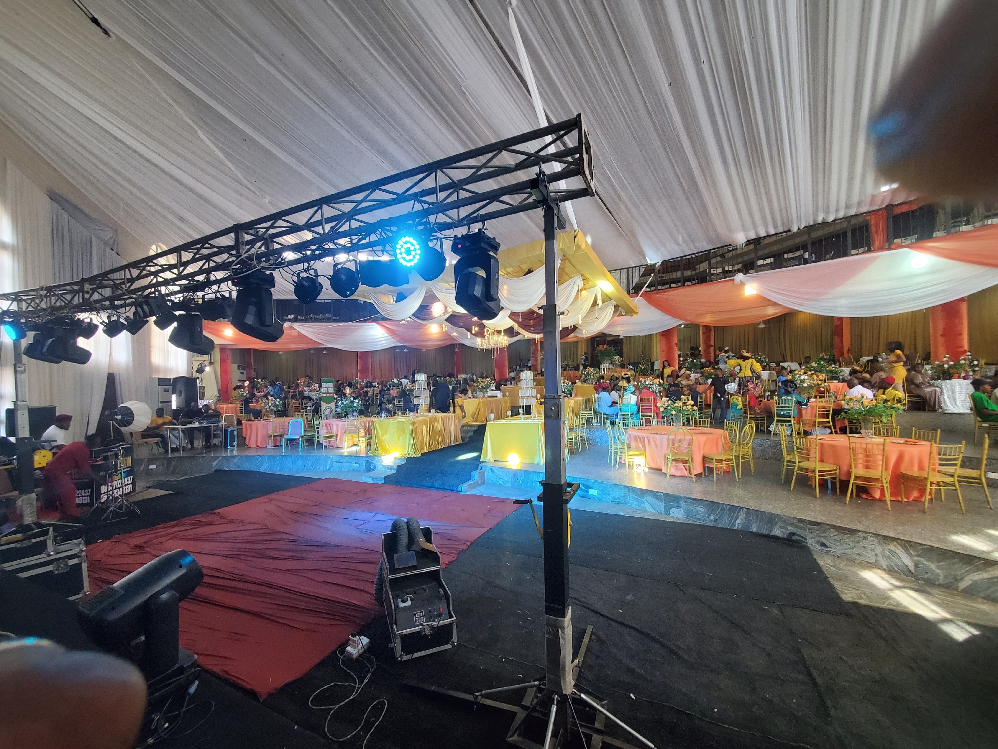 The image depicts the interior of a large event space, likely a reception hall in Onitsha, Nigeria. The space is decorated with draped white and colored fabrics on the ceiling. Multiple round tables are arranged throughout the hall, covered in pastel tablecloths (peach, yellow), and surrounded by golden chairs. People are seated at some of the tables.
In the foreground, stage lighting equipment is visible, including spotlights and a truss system with multiple lights. A stage area is covered in sections of red and blue fabric. A fog machine is located near the stage.
The overall impression is of a large event space set up for a formal occasion.