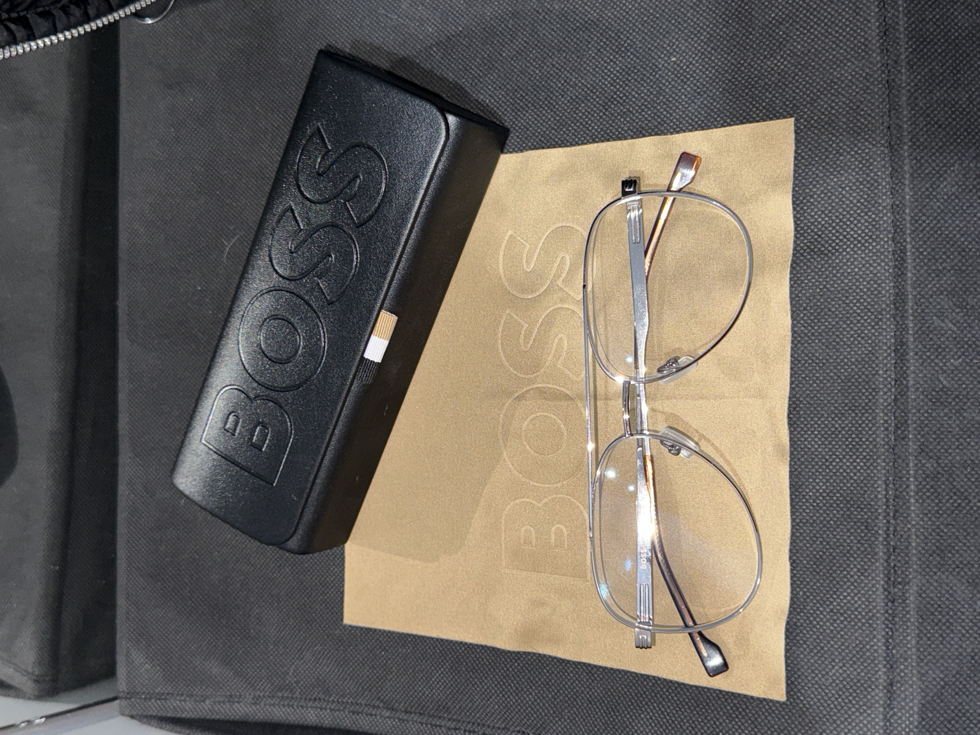 The image shows a pair of Hugo Boss glasses lying on a light brown Hugo Boss cleaning cloth next to a black Hugo Boss glasses case on a dark gray fabric surface. The glasses are silver-rimmed. The case is embossed with the word "BOSS". The cloth is also embossed with the word "BOSS", in a lighter shade than the cloth. The background fabric appears to have a textured, patterned surface. There is no information related to the city of Stara Zagora, Bulgaria.