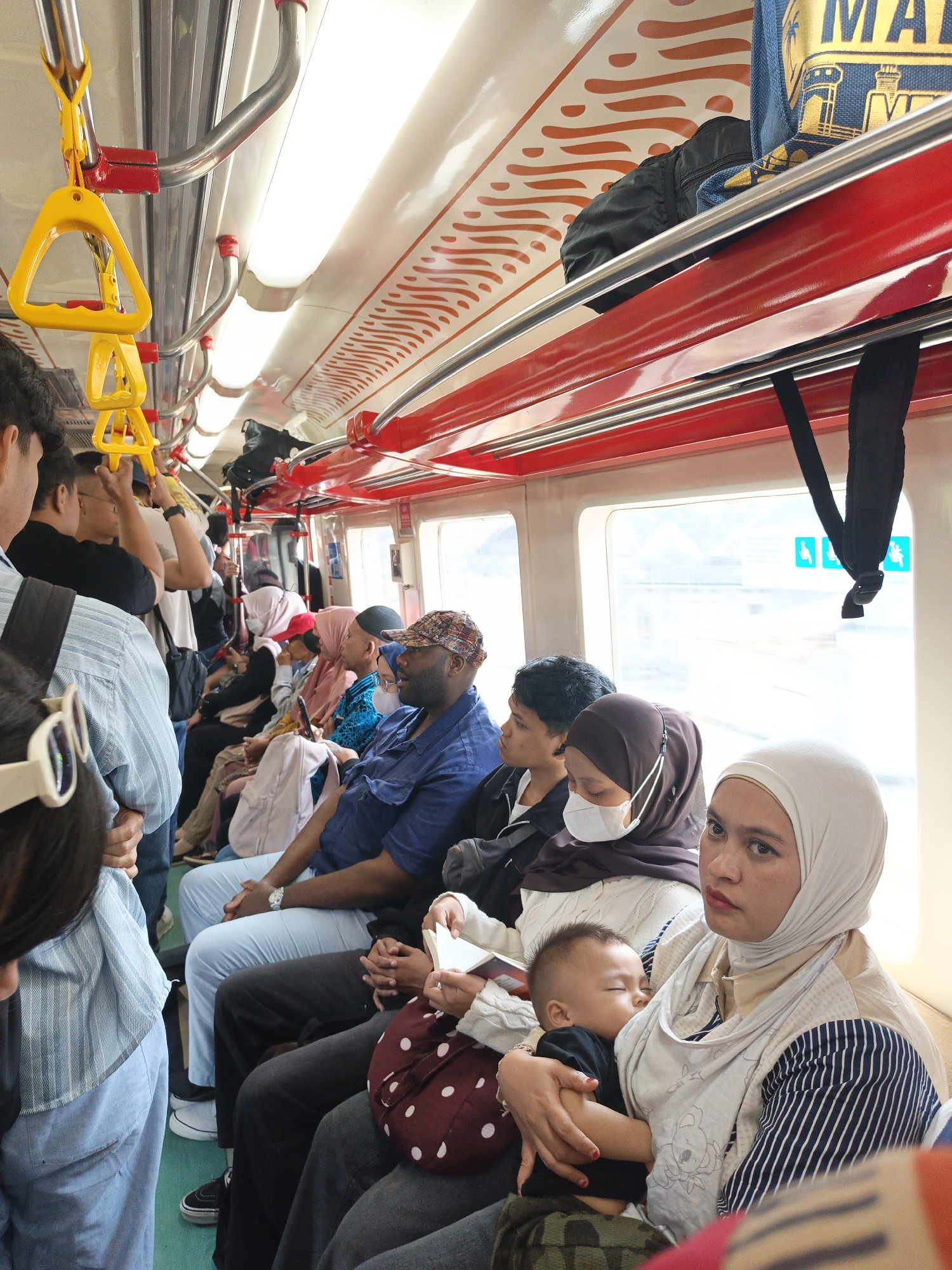 The image depicts the interior of a crowded public transportation vehicle, likely a train or bus, filled with passengers. The passengers are of various ages and genders, many wearing headscarves and modest clothing. Some are seated, while others appear to be standing or holding onto overhead handles, indicated by yellow and red plastic grips visible in the upper left. The interior features red accents on the walls and ceiling, with bright fluorescent lighting. A young child is being held by a woman who appears to be breastfeeding. The environment suggests a warm climate, as indicated by the attire of the passengers. The presence of a sign with what looks like wheelchair accessibility icons is visible through the window. While the location of Pokak, Indonesia is mentioned, the visual elements within the image do not provide specific identifiers to confirm this exact location, beyond it being a public transit setting.