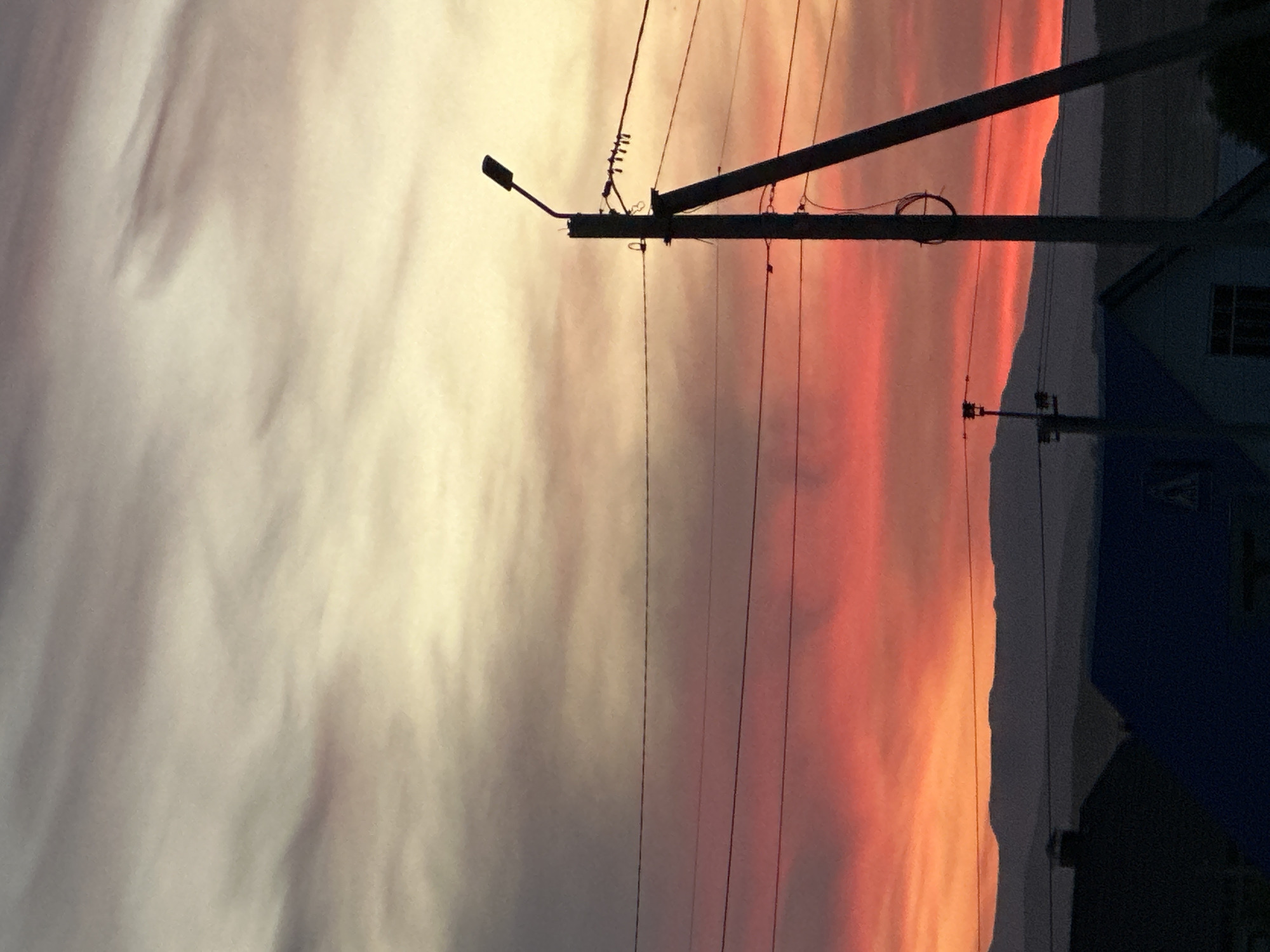 The image captures a scene at dusk or dawn, featuring utility poles and wires against a colorful sky. The sky transitions from pale yellow and gray on the left to orange and red on the right, suggesting a sunrise or sunset. A silhouetted building with a window is visible in the right foreground. Utility poles with attached wires are a prominent feature, adding a structured element to the natural backdrop. The location appears to be in an urban or suburban setting, though the specific city and country are unknown.