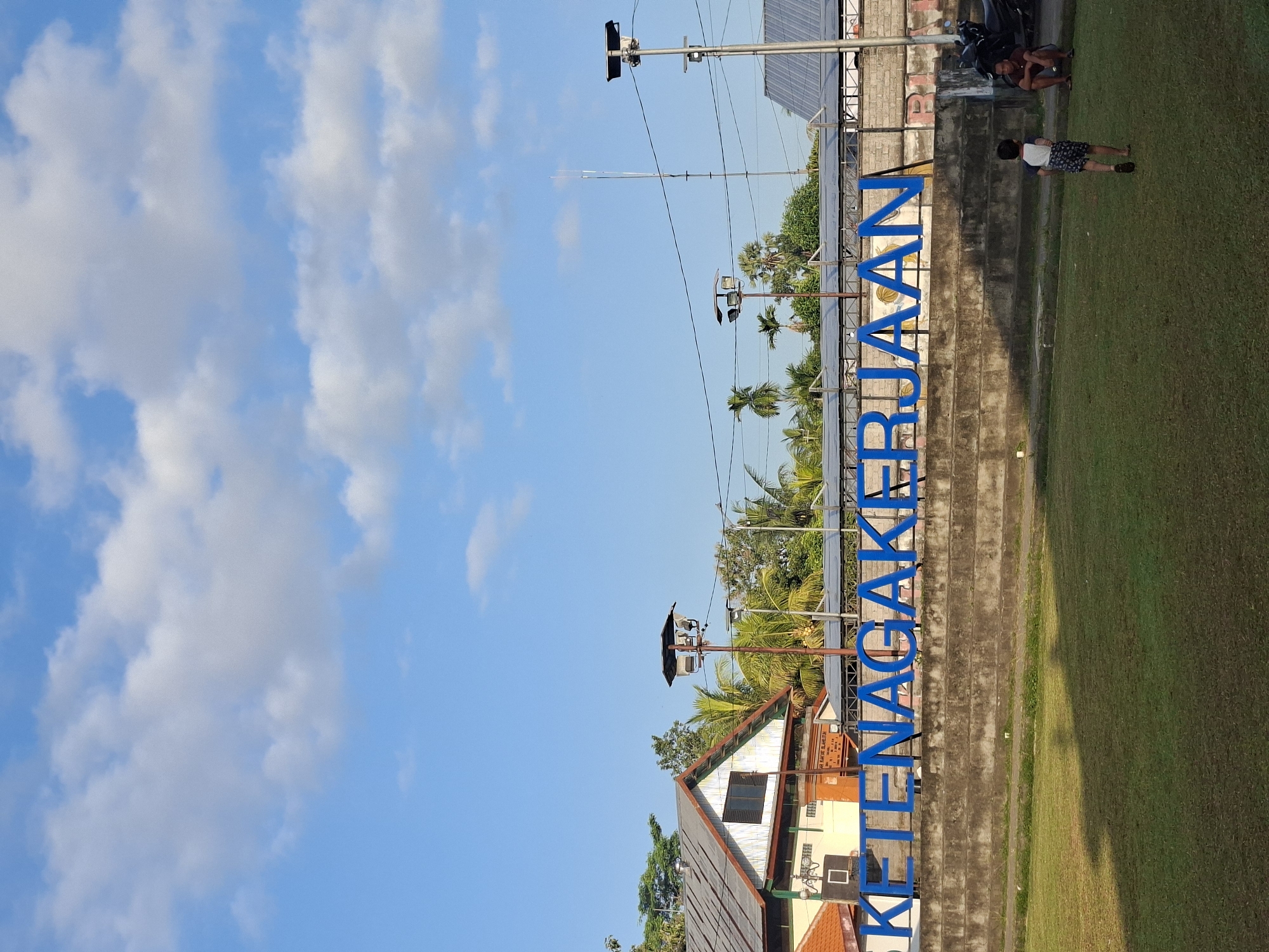 In Blahbatuh, Indonesia, an outdoor scene unfolds under a bright blue sky with scattered white clouds. A large blue sign featuring the words "KETENAGAKERJAAN" is prominently displayed. The sign is positioned behind a tiered concrete structure that overlooks a grassy field. On the field, a young child is walking away from the camera, and an adult male is seated on the ground to the right, near a motorcycle. Buildings with tiled roofs and palm trees are visible in the background, suggesting a tropical or subtropical environment. Streetlights are also present, indicating the area is equipped for nighttime use. The overall impression is one of a public space, possibly a park or community area, in the town of Blahbatuh.