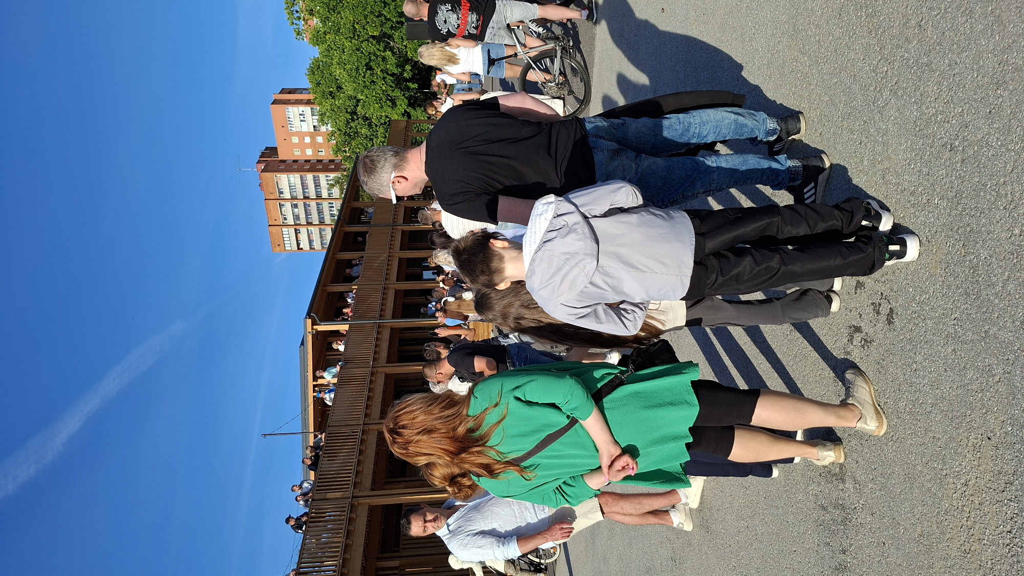 The image depicts a daytime outdoor scene with a group of people standing on a paved area in front of a wooden structure. The building features an upper level with a balcony or deck area where several individuals are gathered, looking outwards. The sky is clear and blue with a faint contrail visible. In the background, multi-story brick buildings and lush green trees can be seen, suggesting an urban environment.
In the foreground, several people are visible from the waist down. A woman with long red hair wearing a green top and black leggings is standing with her hands clasped behind her back. To her left, a man in a light blue collared shirt and beige shorts is looking towards the left. In front of the woman in green, a young boy in a grey hooded jacket and black pants is standing. To the right of the boy, a man in a black t-shirt and blue jeans is facing away from the camera. A bicycle is visible on the right side of the image, near a woman in a white top and denim shorts. The pavement is a mixture of asphalt and gravel, with distinct shadows cast by the people and the building. The overall impression is that of a public gathering or event.
The presence of similar multi-story brick buildings and the general architectural style of the wooden structure is consistent with areas in Tallinn, Estonia, particularly those near the coast or in regenerated urban districts. The building in the background resembles residential blocks found in various parts of the city.