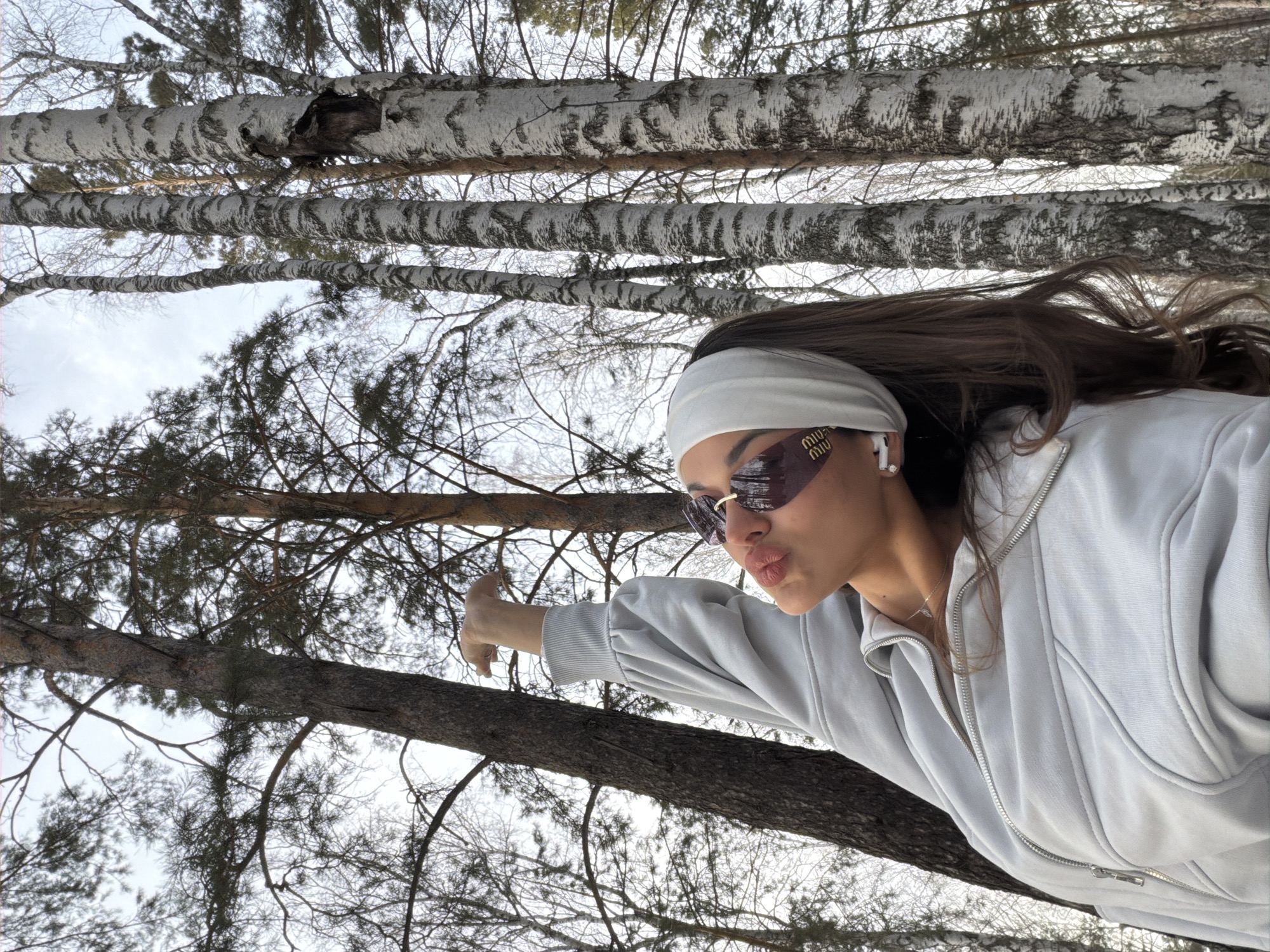 A young woman wearing a light grey hoodie, white headband, and sunglasses is shown in a ground-level perspective, looking upward into a forest of bare trees. She is pointing to the left of the frame. The trees are mainly birches with some evergreens visible in the background. The sky is overcast. The location is likely a park or wooded area in Krasnoyarsk, Russia.