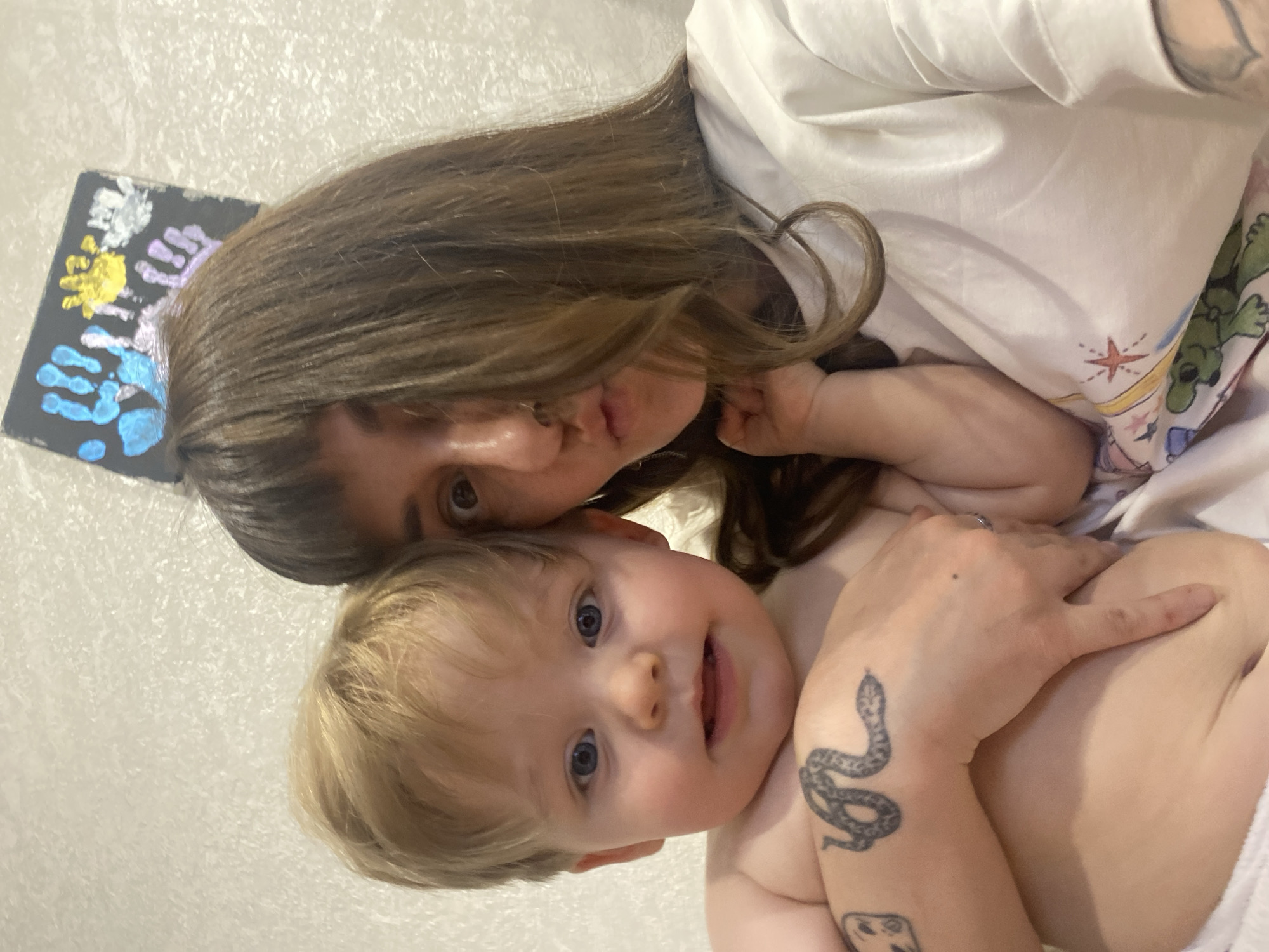 A person with long brown hair and a light complexion is holding a blond-haired toddler with blue eyes. The person is wearing a white shirt and has tattoos on their arm. To the left of the person is a black board with colored handprints on it. The background appears to be a light-colored wall with a slight texture. There is no information to indicate the location is in Votkinsk, Russia.