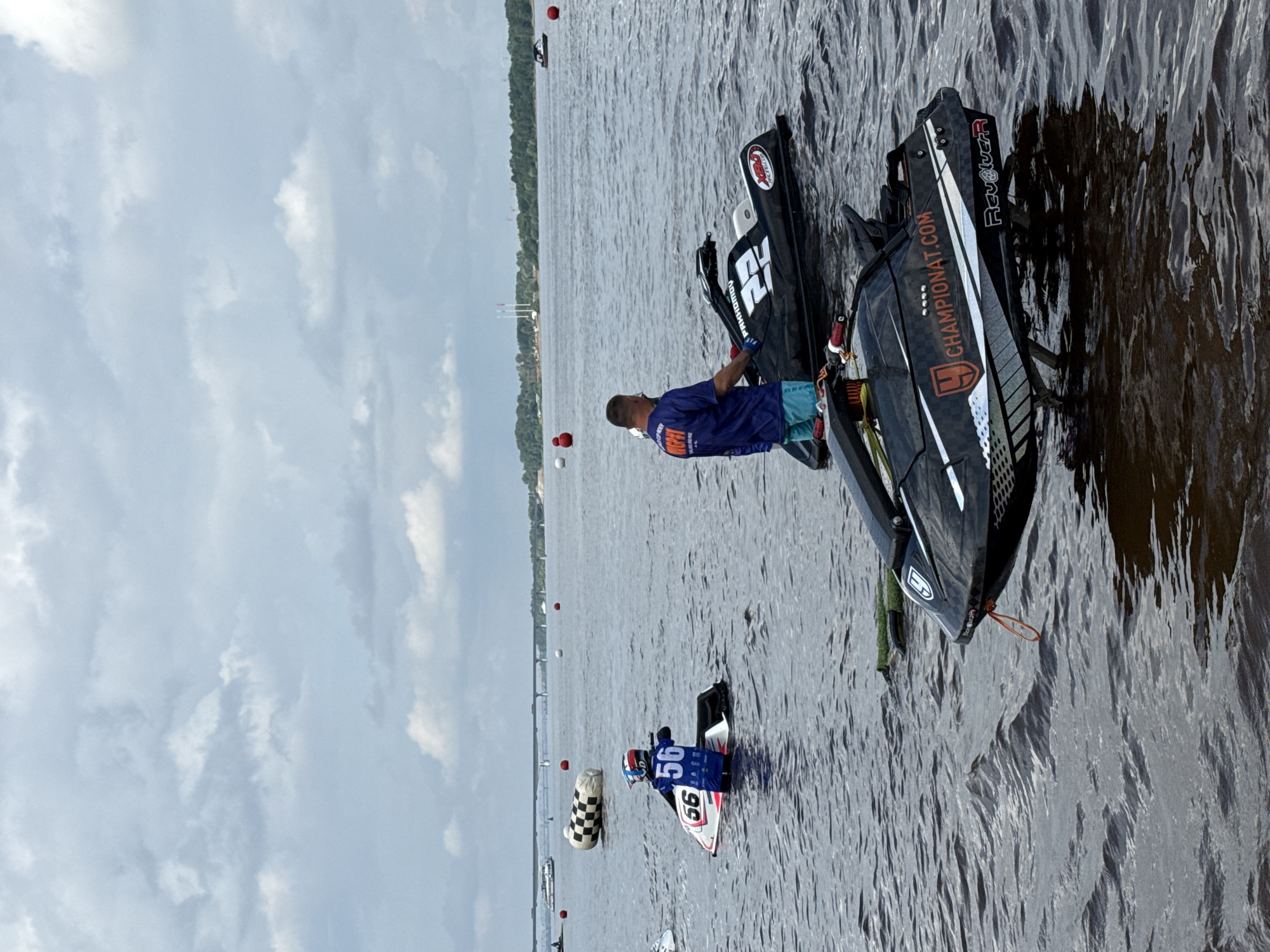 An outdoor, full shot captures a jet ski race event on a cloudy day, with a city skyline and bridge in the background. The water is dark and choppy, with several jet skis scattered across the surface. In the foreground, two jet skis are partially submerged, with one jet ski in focus, a black and white racing model with the text "CHAMPIONAT.COM" visible on its side. A person in a blue t-shirt and turquoise shorts stands next to this jet ski, facing away from the camera. To the left, another jet ski, pink and white, is occupied by a person wearing a blue racing suit and helmet with the number "56" prominently displayed. In the middle ground, a checkered buoy is floating on the water, and further back, a bridge crosses the river. The sky is filled with dramatic, grey clouds, suggesting an impending storm. The location appears to be the Kama River in Perm, Russia, given the distinctive cityscape and infrastructure visible.