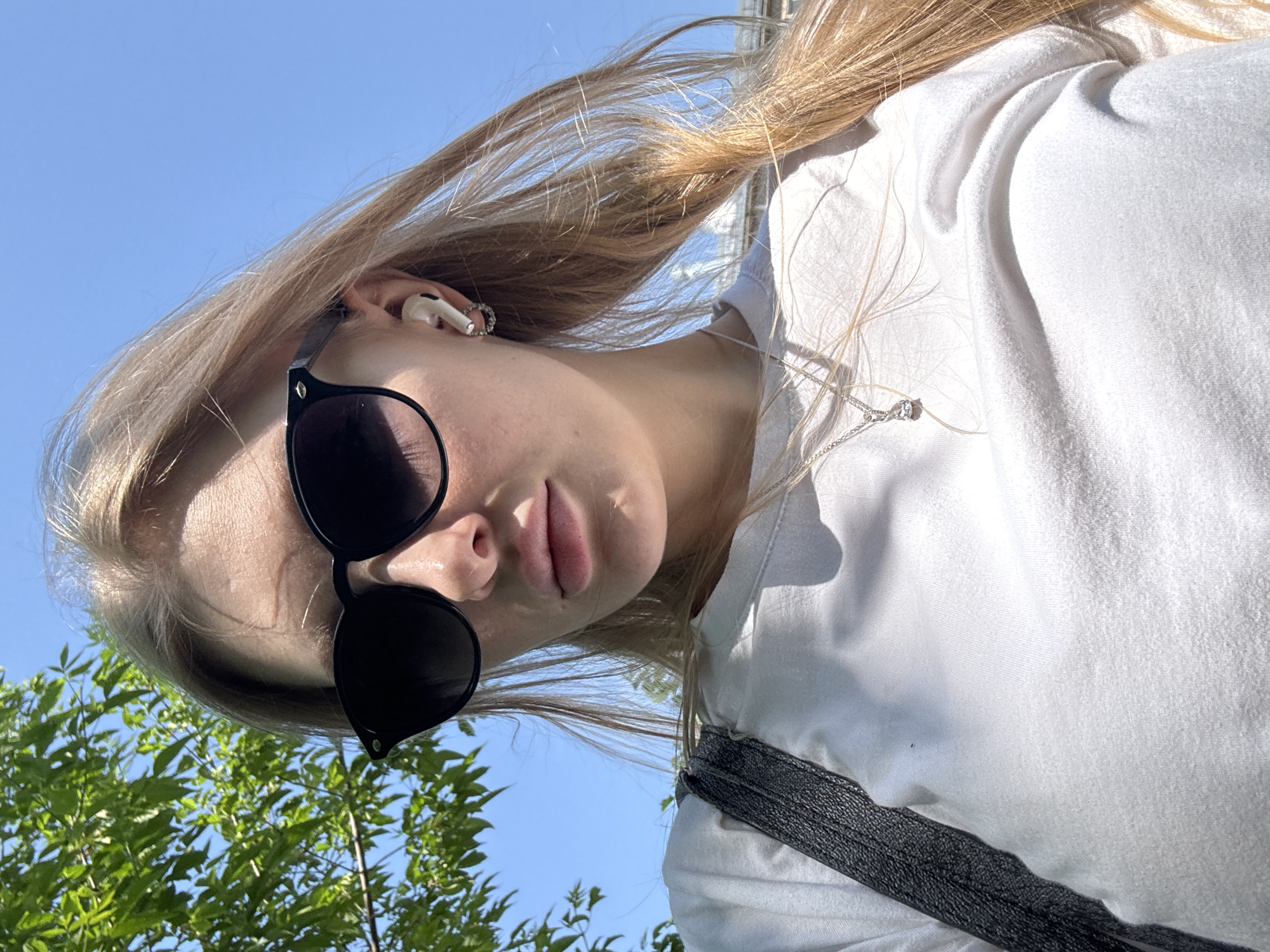 A low-angle shot depicts a fair-skinned young woman with blonde hair wearing black sunglasses and a white t-shirt. An earbud is visible in her left ear. A silver necklace adorns her neck. The background shows a clear blue sky and green tree foliage. Based on the available information, I cannot determine if this image was taken in Perm, Russia.