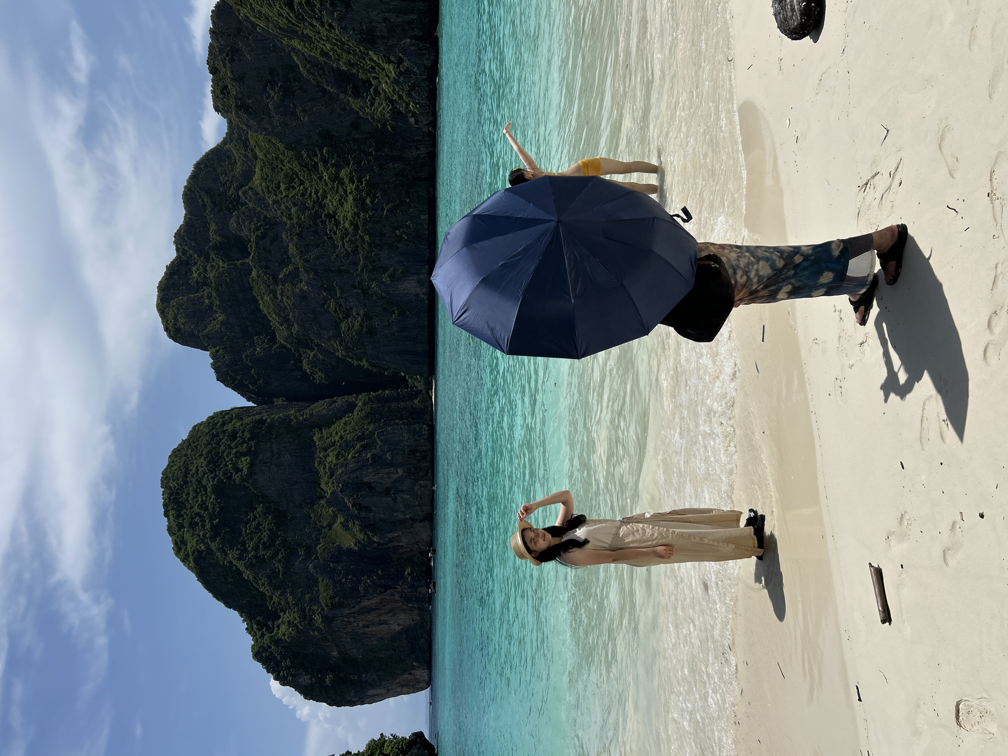A wide shot captures a tropical beach scene in Ao Nang, Thailand, under a partly cloudy blue sky. In the foreground, a person stands with their back to the camera, holding a large, dark blue umbrella. Their lower body is visible, wearing patterned trousers and dark sandals. The person casts a long shadow on the white sand.
To the left of the foreground figure, a young woman in a light-colored, sleeveless dress and a straw hat stands facing the camera, holding the brim of her hat with her right hand. She is positioned at the edge of the shallow, turquoise water.
Further back in the water, another young woman, partially obscured by the umbrella, is visible in a yellow bikini. She has her left arm outstretched.
The beach is composed of fine, light-colored sand with some scattered debris, including a piece of driftwood in the middle ground and a dark, rough object on the right. Footprints are visible in the sand.
The ocean is a striking turquoise color, with gentle waves lapping at the shore. In the background, majestic, tree-covered limestone karst islands rise dramatically from the water. Several longtail boats are visible in the distance on the water. The overall impression is a serene tropical paradise.