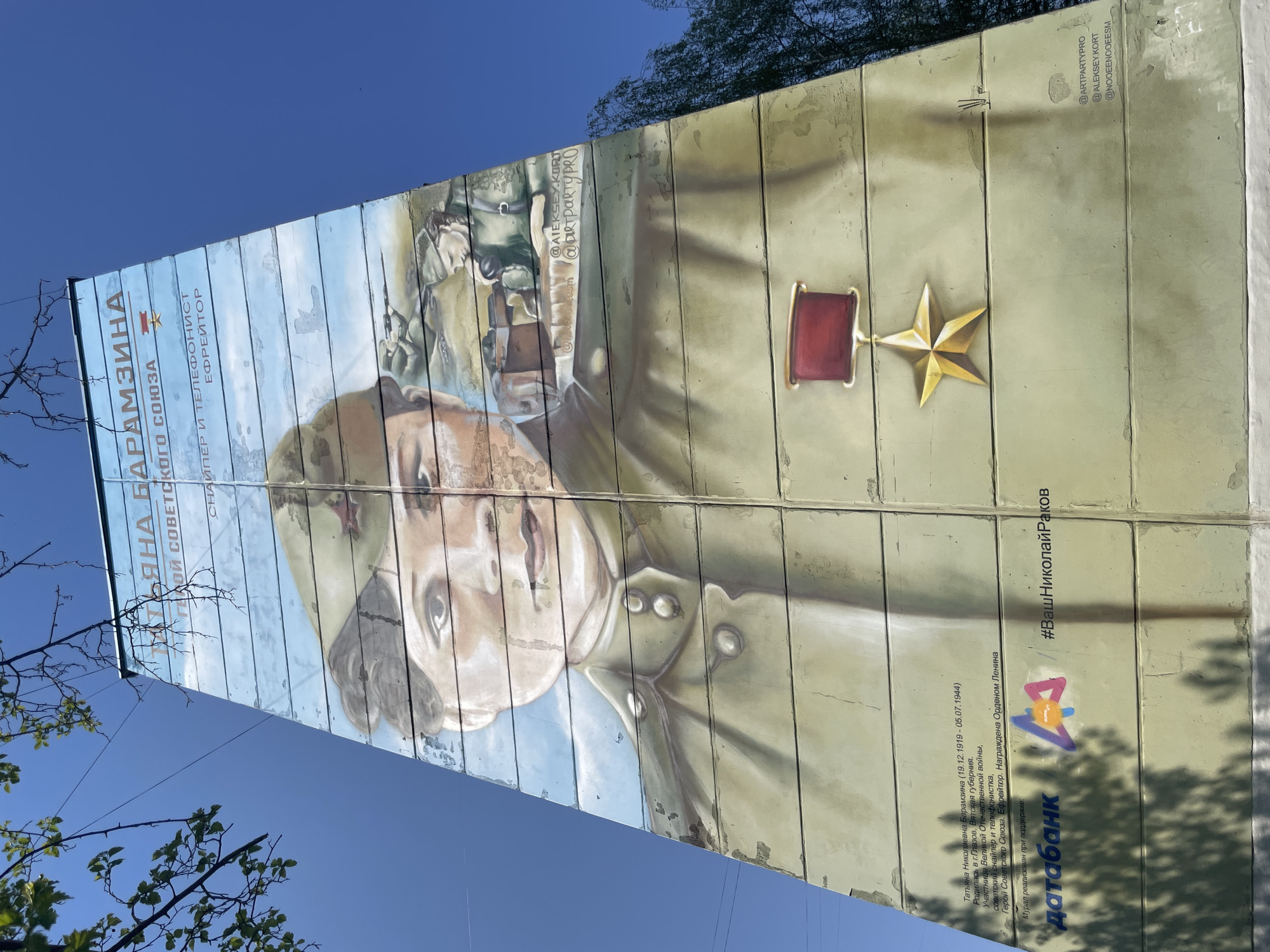 The image shows a mural on a multi-section wall in Izhevsk, Russia. The mural depicts Tatiana Nikolaevna Baramzina, a Soviet sniper and telephone operator during World War II who was awarded the Order of Lenin and who died in 1944.
Baramzina is depicted in a light-colored military uniform, with a red-and-gold Soviet medal on her right. In the background, an image of soldiers involved in a battle scene can be seen. Text on the mural includes her name, rank and details about her life, as well as the hashtag "#ВашНиколайРаков".
The lower part of the mural features the logo of "Датабанк" (Databank) along with credits for the artists with their social media usernames. The mural is painted on a textured surface which appears to be segmented. Parts of trees are visible in the foreground and background. The sky is clear and blue.