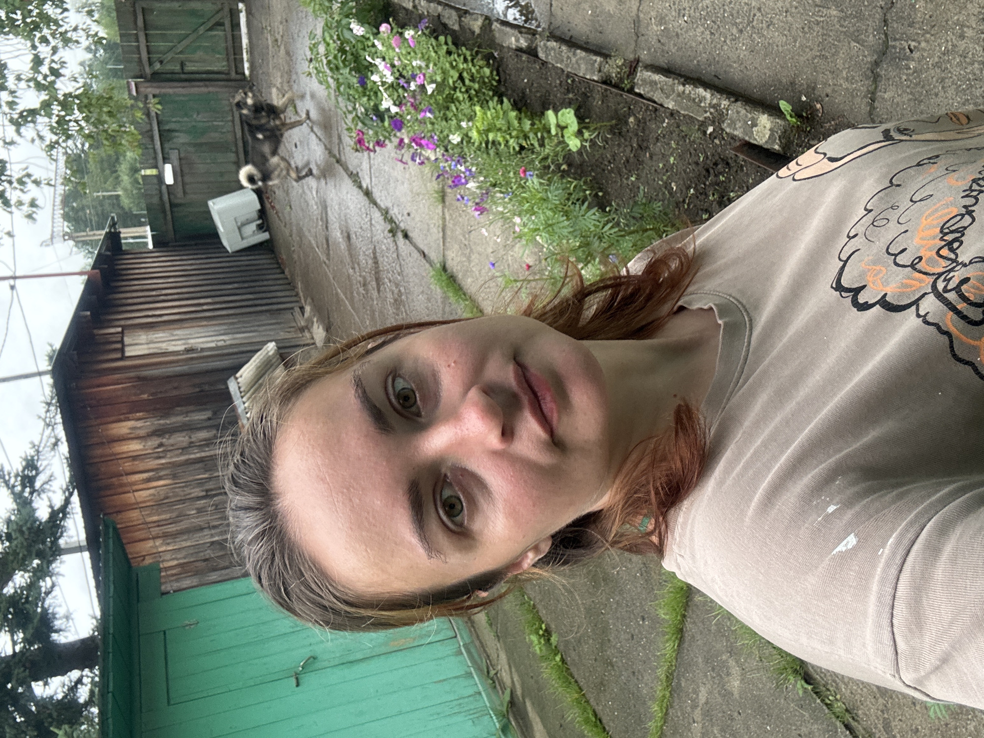 The image depicts a selfie taken outdoors, presumably in a yard or rural setting. A woman with light brown hair and green eyes is the central subject, looking directly at the camera. She is wearing a light brown t-shirt with a graphic print on the front, and there are some white paint-like splatters on the left shoulder area.
In the background, there is a weathered wooden structure, possibly a shed or outbuilding, with vertical wooden planks. To the left of this structure is a bright green door. A paved pathway runs through the scene, and along the right side of the pathway is a flower bed with various colorful flowers, including purple, pink, and white varieties, as well as green foliage.
Further in the background, a wooden fence with green paint encloses the area. A dog, appearing to be a medium-sized breed with a mix of brown and black fur and a bushy tail, is visible in the middle ground, walking on the wet pavement. Behind the dog, a white rectangular object, possibly a piece of discarded appliance, rests on the ground. The ground appears wet, suggesting recent rain. Overhead, tree branches and foliage are visible, with a cloudy sky in the distance. The overall impression is a candid shot taken in a somewhat unkempt, possibly rustic environment.