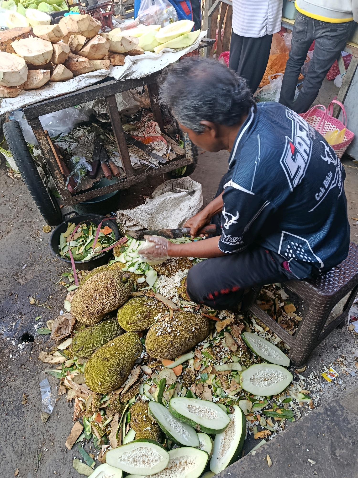 In Gondanglegi, Indonesia, a man in a black and blue shirt is sitting on a plastic stool, peeling jackfruit. The scene appears to be an outdoor market, with a cart filled with fruits and vegetables in the background. The ground is littered with discarded fruit peels, seeds, and other organic matter. There are several whole and cut jackfruits, as well as cut papayas scattered around the man. A black bucket containing more fruit and vegetable scraps is also visible. Two pink woven baskets are placed near a stall in the background. The overall atmosphere suggests a busy, perhaps slightly unkempt, market environment.