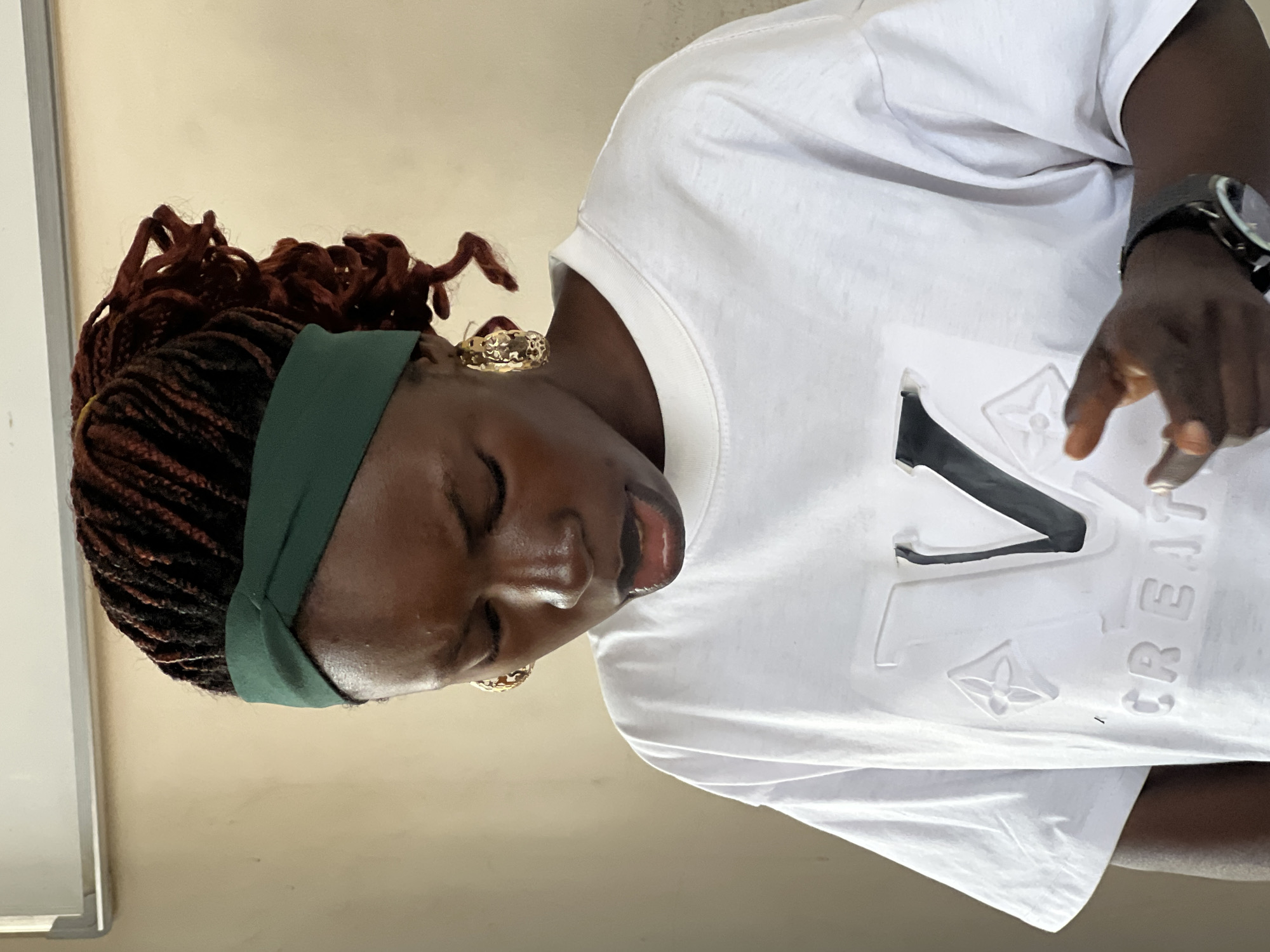 A young Black woman with dark skin, styled in red-brown box braids and wearing a green headband, is captured from the chest up, looking downwards and to her left. Her eyes are closed, and her mouth is open as if speaking. She is adorned with large, textured gold hoop earrings. She wears a plain white t-shirt with a prominent, embossed design featuring a large "V" intertwined with floral-like shapes and the word "CREATIVE" below it. Her right hand, with painted fingernails, is extended forward, fingers slightly curled. A dark wristwatch is visible on her left wrist. The background consists of a plain, beige wall with a section of a white whiteboard visible at the top left, suggesting an indoor setting, possibly a classroom or office. The lighting suggests the image was taken indoors with artificial light. The context of Jos, Nigeria, is not directly discernible from the visual elements of the image itself.