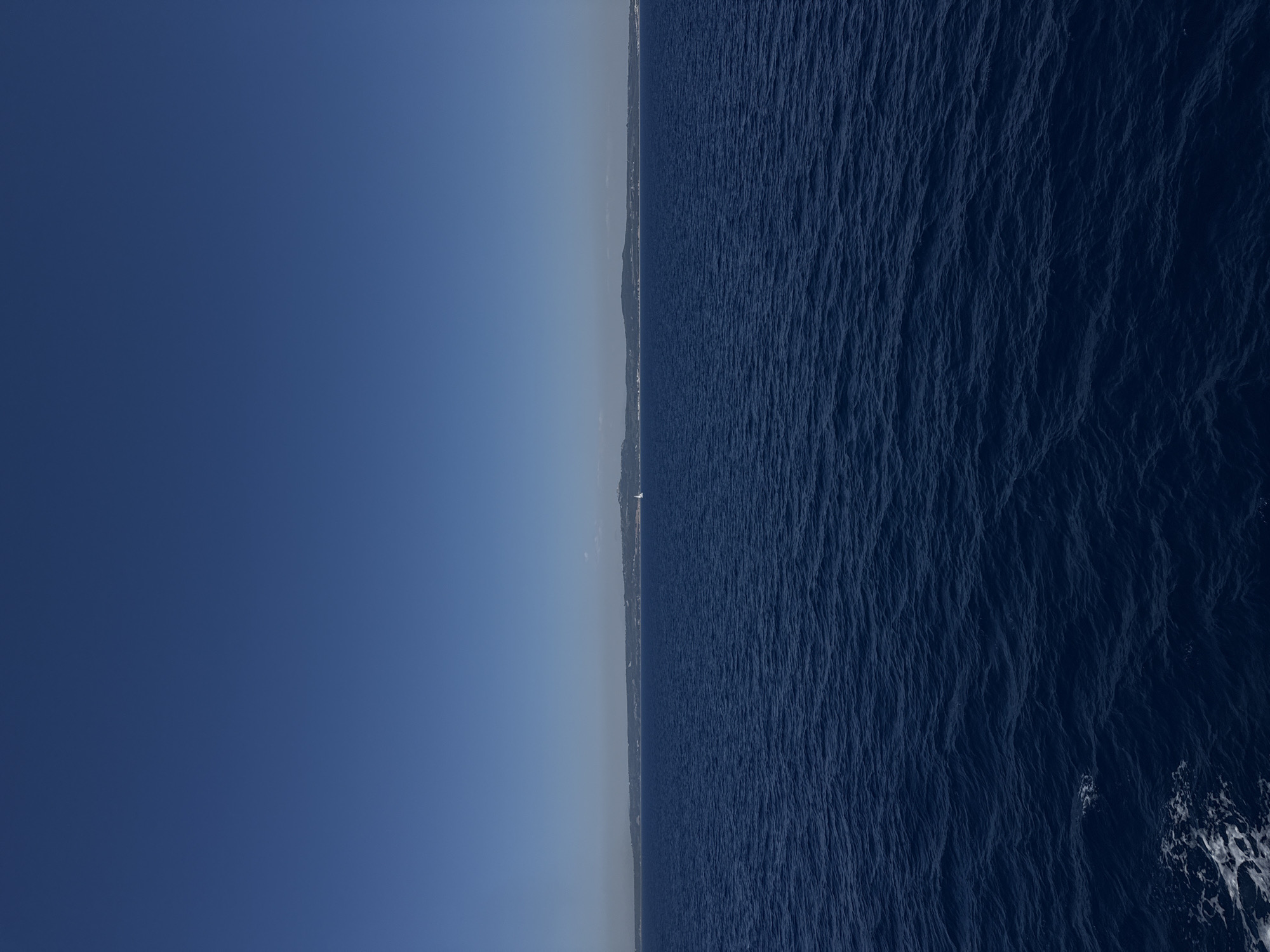 This is an outdoor, eye-level, horizontal, medium-distance shot of the sea and sky. The scene is dominated by the deep blue of the ocean in the foreground, with gentle waves creating a textured surface. In the middle ground, a distant, hazy coastline stretches across the horizon, with subtle hints of green vegetation and white structures suggesting a town or settlement. A single, small white sailboat is visible on the water, to the right of the image. The upper portion of the image is filled with a clear, uninterrupted expanse of deep blue sky. The overall impression is one of vastness and tranquility. The location is the sea off the coast of Kalamata, Greece.