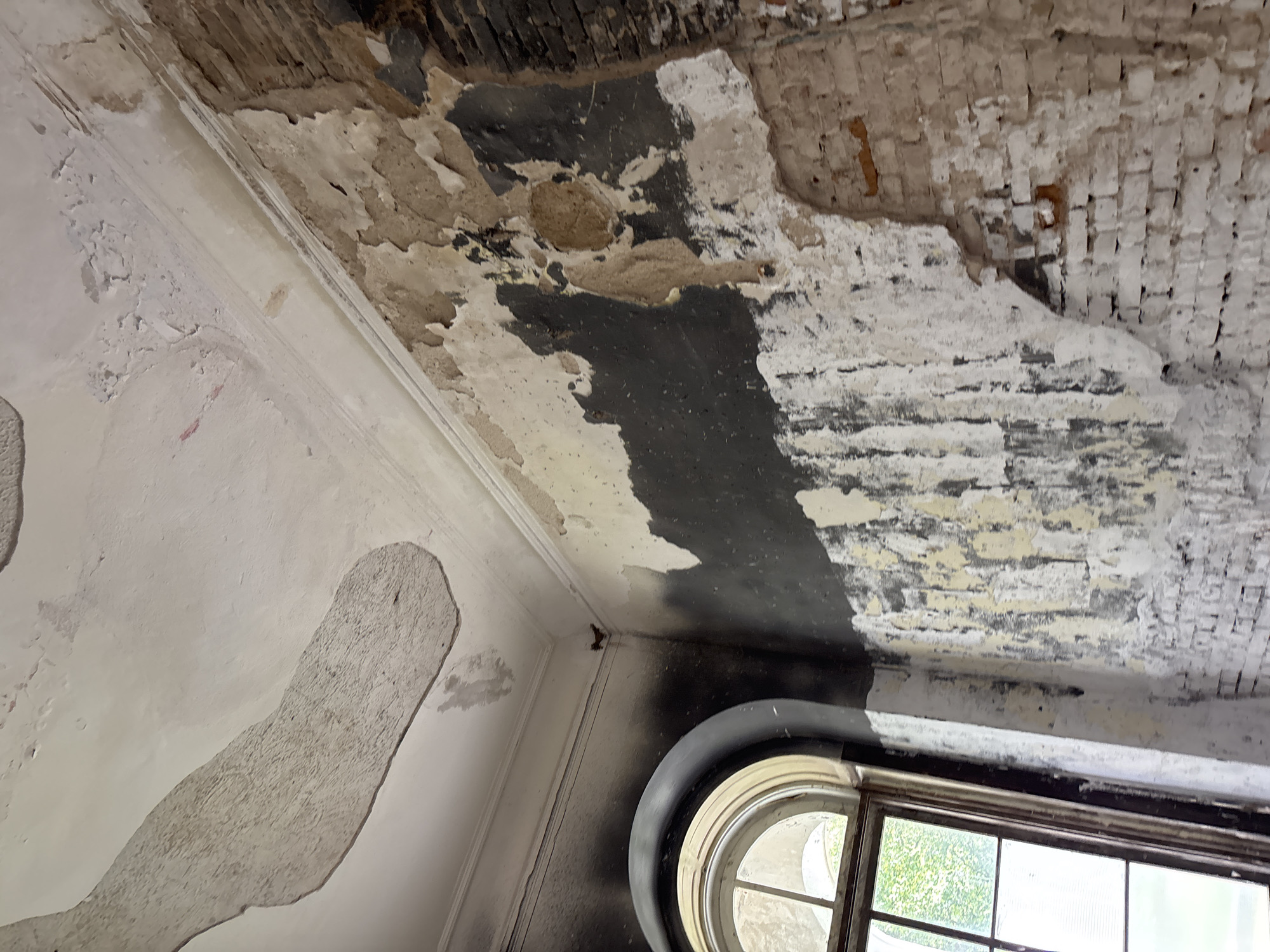 The image depicts the interior of a room in the Zhaludok Synagogue, located in Zhaludok, Belarus. The scene is characterized by significant disrepair, suggesting a state of abandonment or damage. The walls and ceiling show extensive peeling plaster and water damage, with large sections of the ceiling material detached and hanging down. The walls are a mix of exposed brickwork and remnants of plaster, with dark soot-like stains visible, particularly around the arched window on the left. The window itself is a large, multi-paned structure with an arched top, letting in natural light and revealing greenery outside. The overall impression is one of decay and neglect within a historically significant building.