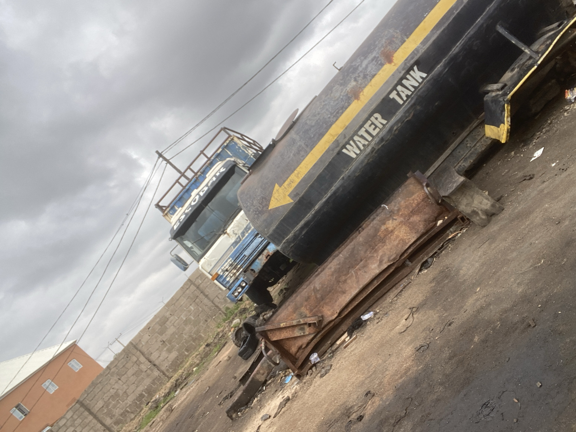 In Gbaza, Nigeria, a blue and white water tank truck is parked on a dirt road. The truck is tilted, with its tank partially obscuring the cabin. The tank is black with a yellow arrow and the words "WATER TANK" written in white. To the left of the truck are several pieces of rusted metal. In the background, a brick wall separates the road from a building with an orange facade and white trim. The sky is overcast with dark clouds.