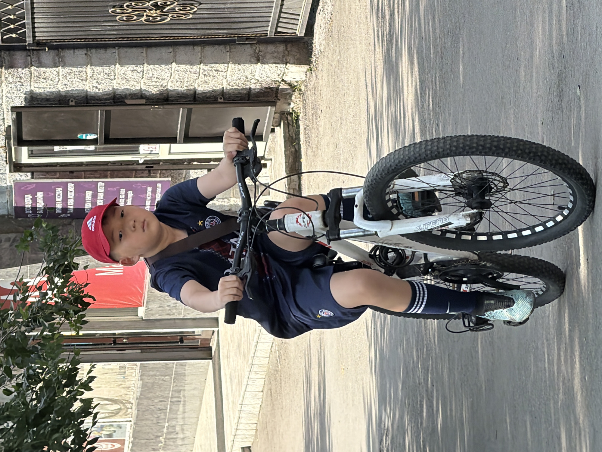 An Asian boy wearing a red baseball cap and a navy blue sports uniform rides a white mountain bike down a paved street. The boy is in the foreground, facing forward and slightly to the left. He has a water bottle attached to the handlebars. In the background, there are buildings and trees. One building has a black metal gate. The sun is shining, casting shadows on the street. The boy's shoes are teal and black, and he is wearing navy blue socks with white stripes. The bicycle has black tires with white sidewall accents. The street is a neutral gray asphalt.