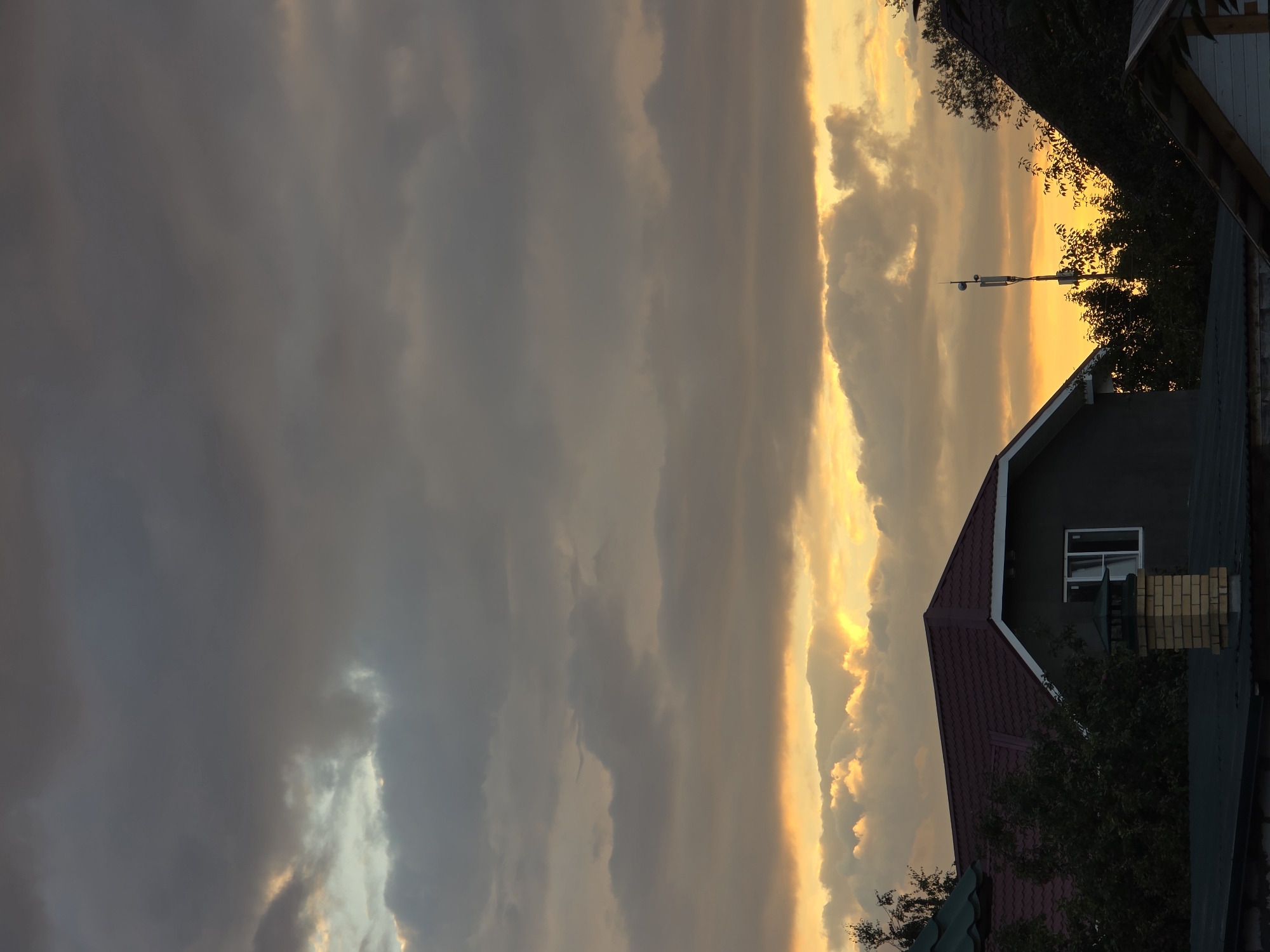 This is a vertical, eye-level shot of a cloudy sky at dusk, with the tops of houses and trees visible in the foreground. The sky is dominated by dramatic, dark gray clouds, with a band of bright, golden-orange light peeking through the lower portion, suggesting the setting sun. Streaks of light emanate from this bright area, illuminating some of the clouds from behind.
In the foreground, the dark red, pitched roof of a house is prominent. To the left, a brick chimney with a green cap rises from another house. Further to the right, the silhouette of a tall telecommunications tower stands against the brightly lit sky. Dense, dark green foliage of trees is visible, partially obscuring some of the structures. The overall impression is of a somewhat overcast but colorful sunset over a residential area. The location is stated as Речицы, Russia.