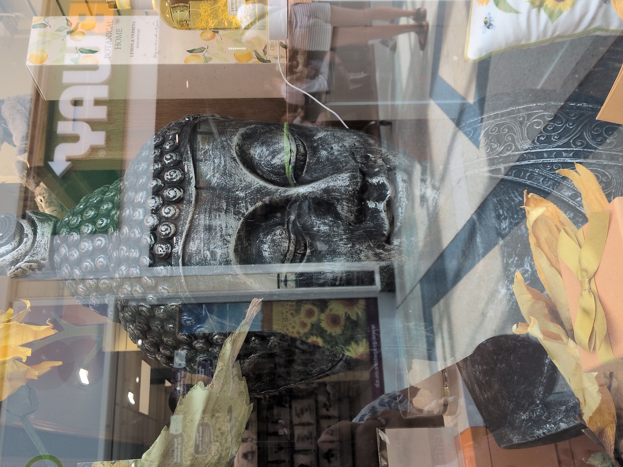 This is a reflective photograph taken through a shop window in Telford, United Kingdom. The main subject is a large, grey, Buddha head statue with a textured, somewhat distressed finish. The statue has a serene expression, with closed eyes and a slight smile. Its head is adorned with a metallic, scaled headdress.

Reflected in the glass are various elements of the environment outside the shop. To the right of the Buddha head, a product box with a "Botanical Home" label and illustrations of lemons and leaves is visible. Next to it, a bottle of liquid, likely a reed diffuser or perfume, with a yellow hue is also present. In the background, a person wearing striped shorts and a white top is partially visible, walking away from the camera. Further reflections show what appear to be shelves stocked with items and a yellow floral motif, possibly sunflowers, on a poster or display.

The shop's interior is dimly lit, with bright rectangular lights visible on the ceiling. The floor appears to be tiled with a dark and light pattern. The overall scene suggests a retail environment, perhaps a gift shop or a home decor store. The presence of the Buddha statue implies a focus on mindfulness, spirituality, or Asian-inspired decor. The reflections create a layered effect, combining the shop's offerings with the external surroundings.