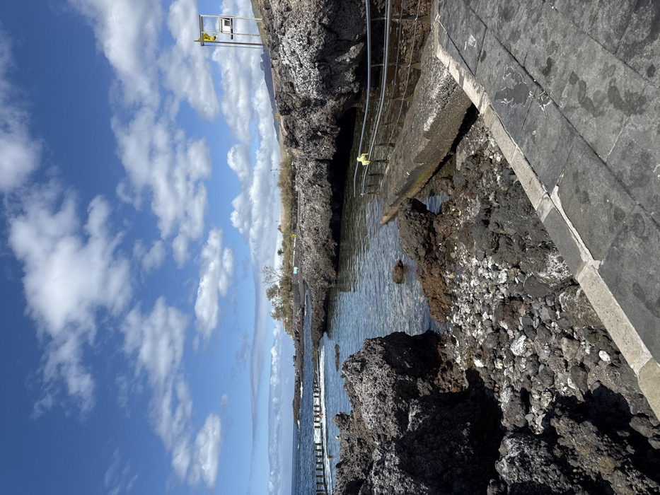 This image depicts a coastal scene in Guía de Isora, Spain, characterized by volcanic rock formations and clear ocean water under a partly cloudy blue sky. In the foreground, a dark grey paved walkway extends along the lower right. Adjacent to this, a concrete ramp with a silver metal handrail leads into a natural pool of transparent, clear water. A yellow object is draped over the handrail. The shoreline is composed of dark, porous volcanic rocks and pebbles, with some lighter-colored debris mixed in.
Further into the scene, the natural pool is surrounded by substantial dark rock formations. In the distance to the left, a section of the ocean is demarcated by a series of vertical wooden posts, forming a natural swimming area. One individual, partially submerged, is visible near these posts. Beyond this, the open sea extends under a bright sky.
On the higher ground to the right, a tall, light-colored metal structure, possibly a signpost or a lifeguard stand, features a yellow flag or cover at its apex. Sparse, low-lying coastal vegetation and distant structures are visible along the coastline. The overall environment suggests a natural, rugged coastal area adapted for recreational use.