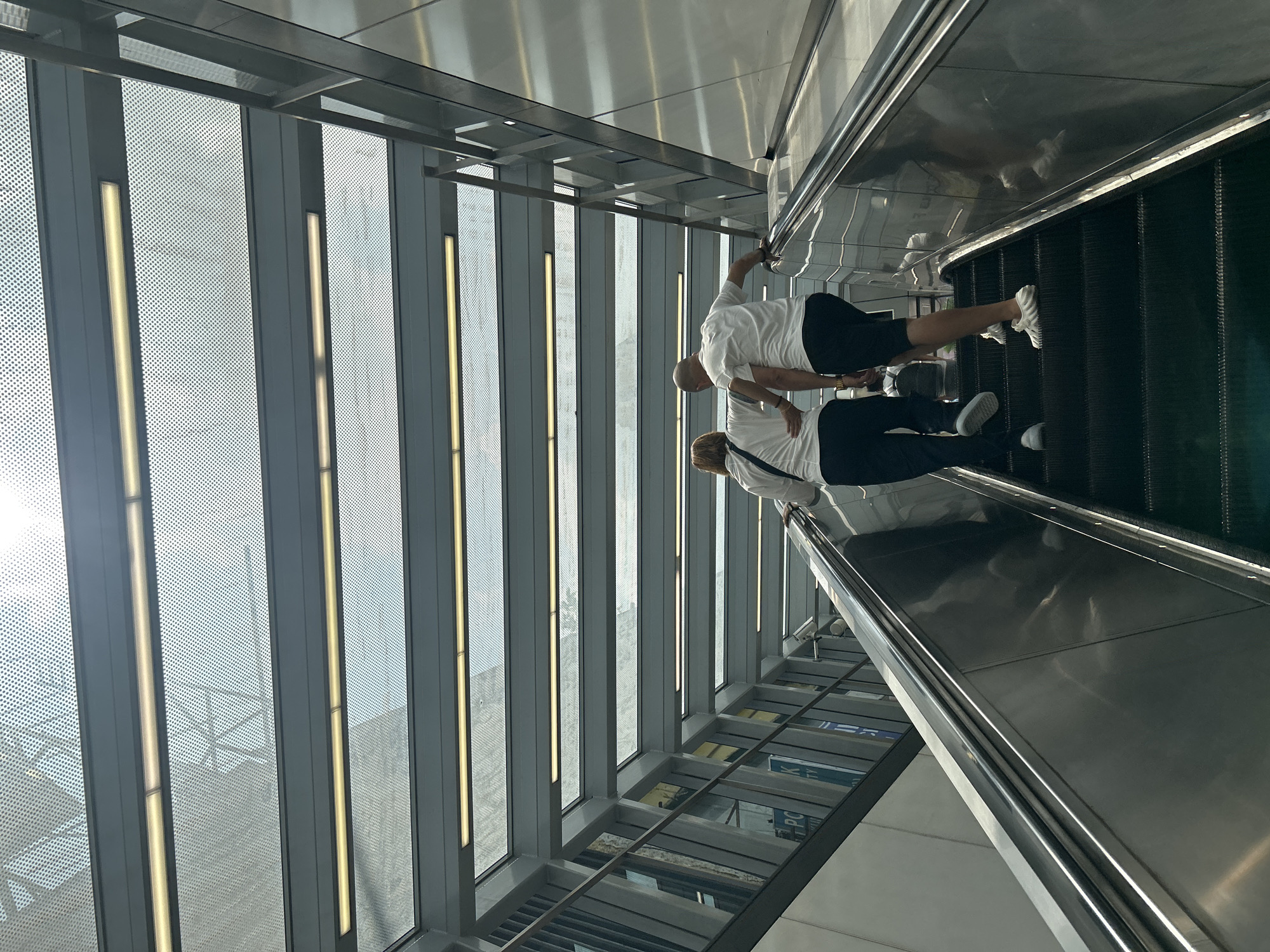 Two individuals, a man and a woman, are ascending an escalator. The woman, on the left, is wearing a white shirt and dark pants. The man, on the right, is also in a white shirt and dark shorts. Both are facing away from the camera and appear to be walking up the escalator. The escalator is made of metal with black steps.
The environment suggests an indoor public space, possibly a modern transit station or a large commercial building, with a glass and metal roof structure overhead. Fluorescent lights are visible along the ceiling. The architecture features a lot of metal and glass elements, with a contemporary design. The overall impression is one of clean, modern lines and an abundance of natural and artificial light. This scene appears to be in Kuala Lumpur, Malaysia, potentially at a location like the KL Gateway Mall or KL Sentral which feature modern architecture with escalators and covered walkways.