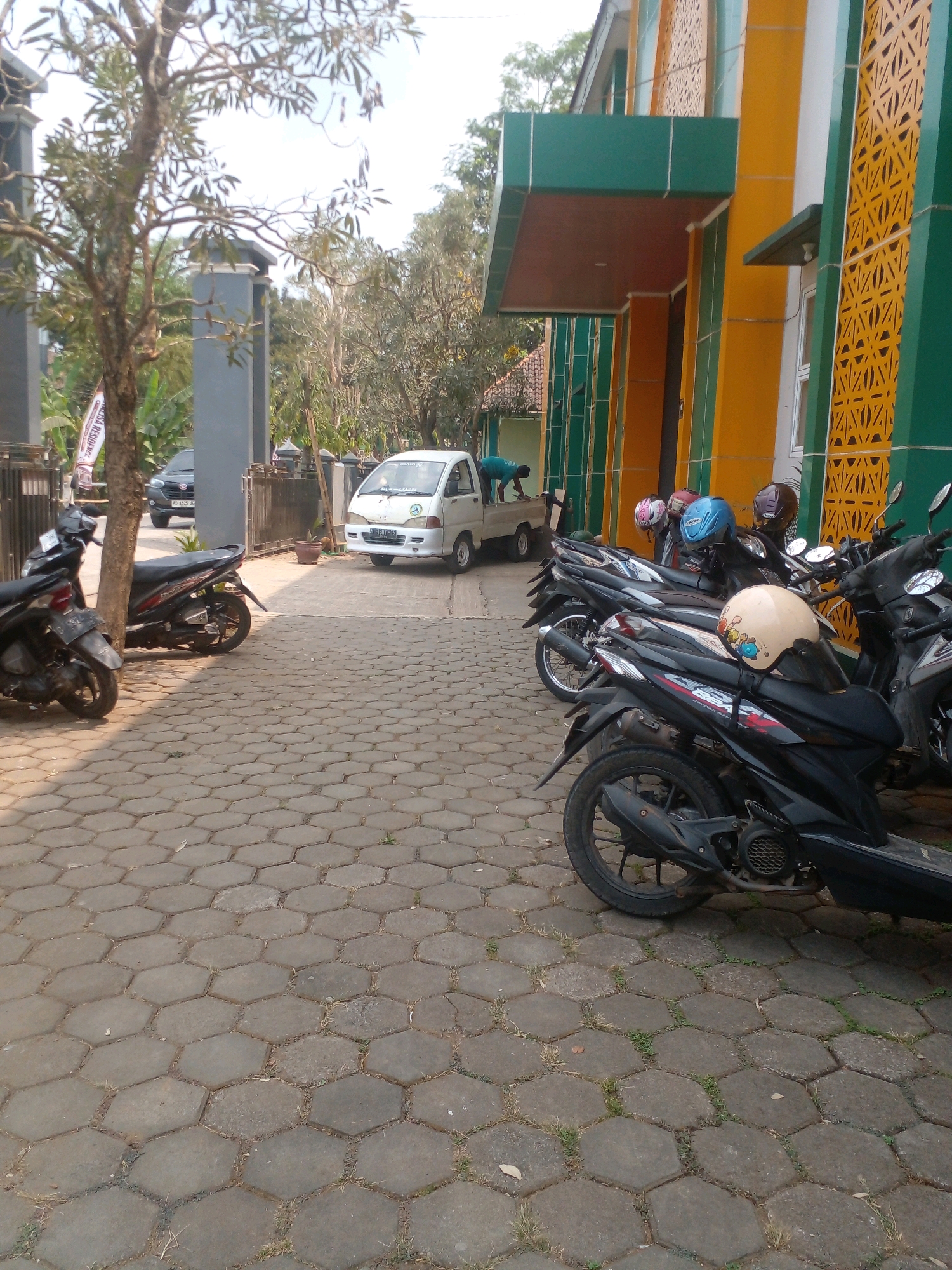 The image depicts an outdoor scene in front of a building with yellow and green architectural features. Several motorcycles are parked on a hexagonal paver stone ground. In the background, a white pickup truck is visible with a person near its rear. To the left, a tree stands next to a fence. The scene appears to be a public area in Banyuputih, Indonesia, possibly a parking lot or entrance to a facility.