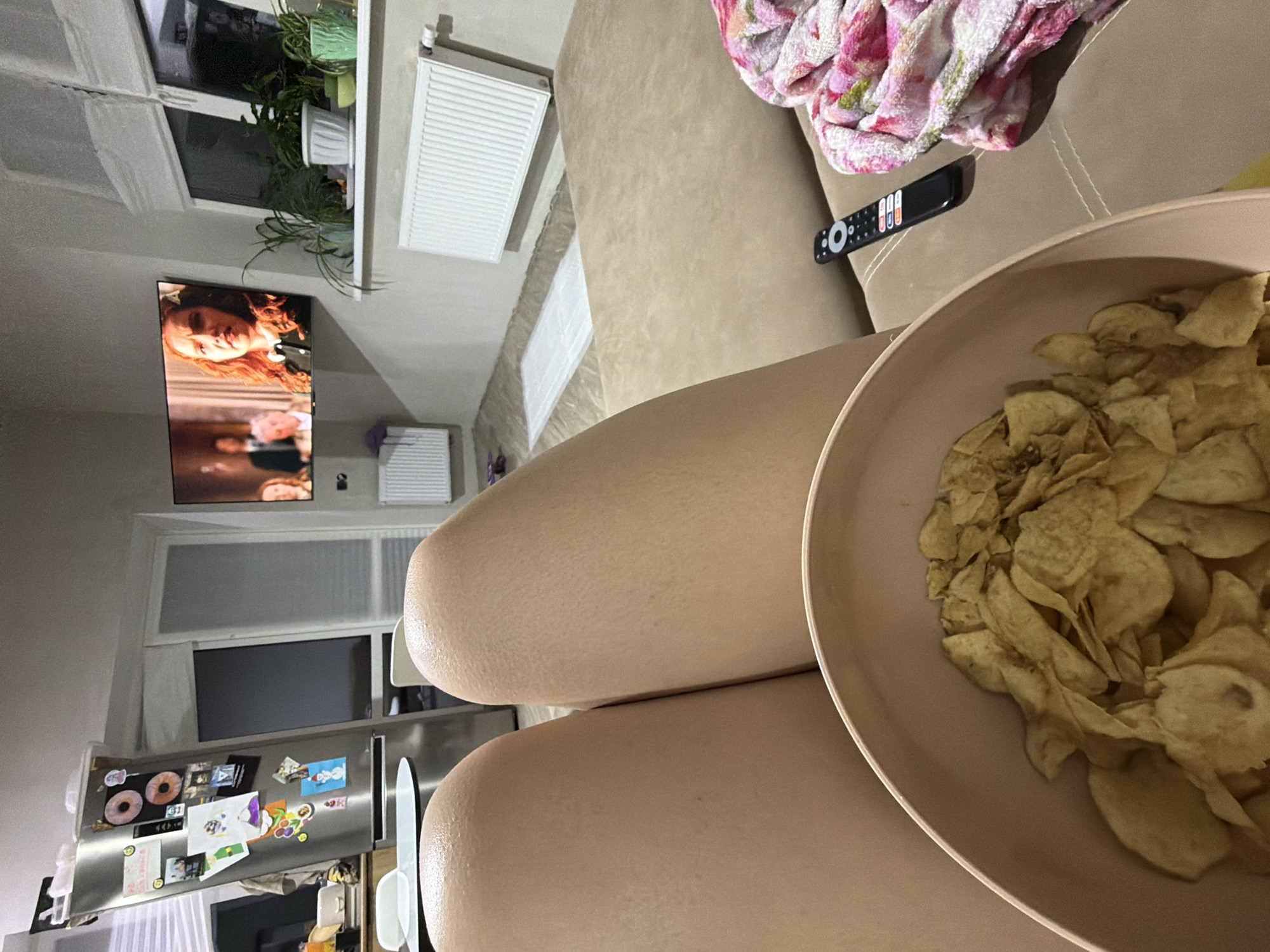 The image shows a person sitting on a beige sofa, likely indoors in an apartment. A large bowl of potato chips is positioned prominently, partially obscuring the person's legs. A TV remote lies nearby on the sofa. In the background, a television screen displays a scene from a show or movie. Through a window, green plants are visible. Further in the background, a refrigerator adorned with magnets and drawings is seen. This suggests the location is a living room or a similar space inside an apartment. The city is unknown.