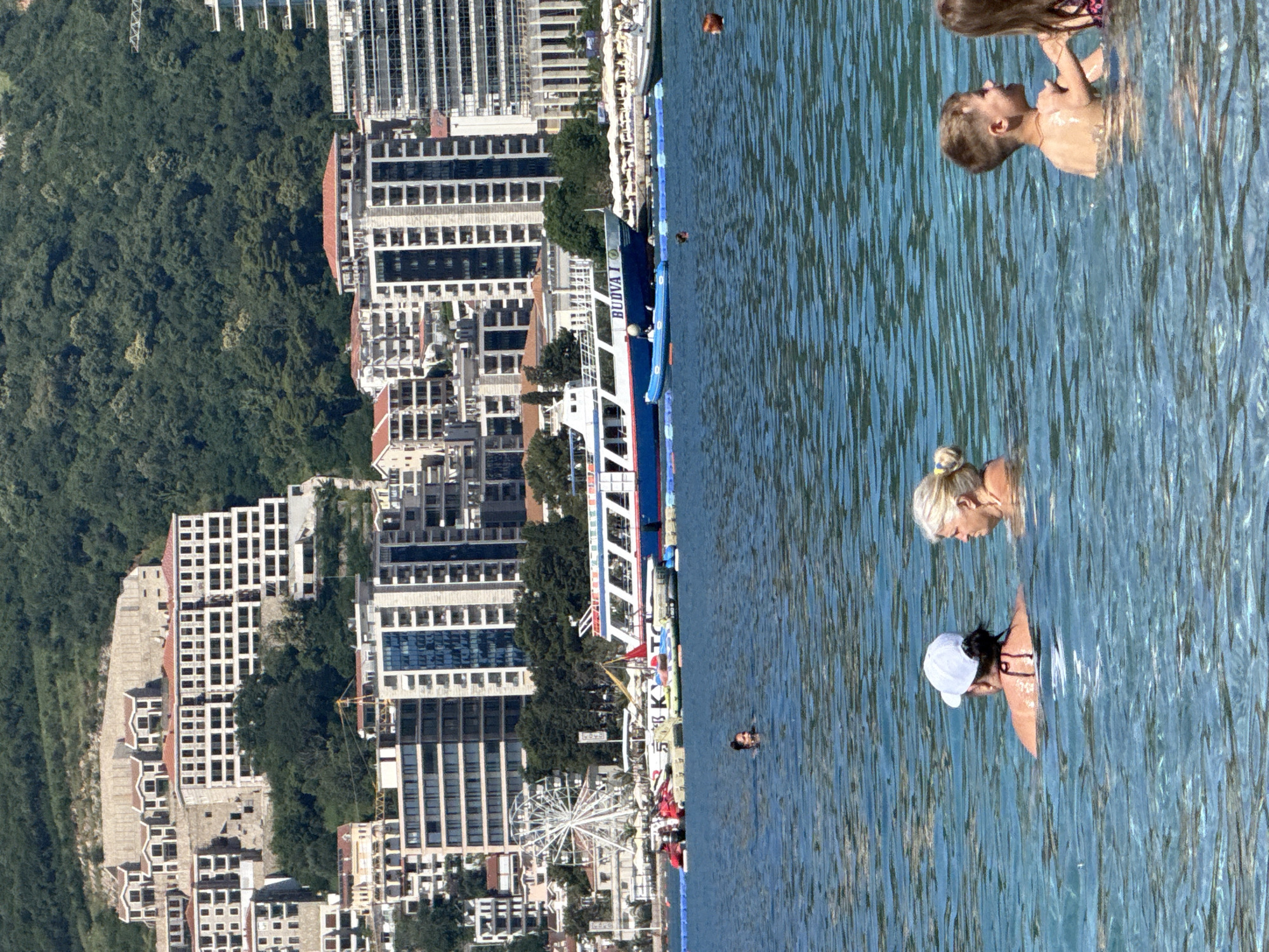 This detailed image depicts a bustling coastal scene in Boreti, Montenegro, under clear blue skies. The foreground features calm, turquoise water where four individuals are swimming: two adults (one wearing a white cap, the other with blonde hair) and two children (a boy and a girl) are visible in the lower right. A fifth person's head is partially visible further back in the water.
In the midground, a blue buoy line stretches across the water, separating the swimming area from a small pier or floating dock. Moored near this structure are several jet skis and inflatable water toys. A large blue and white passenger boat, named "BUDVA I," is prominently visible, either docked or anchored. Beyond the pier, a white Ferris wheel stands on the shoreline.
The background is dominated by dense urban development rising up a steep, heavily forested hillside. Numerous multi-story buildings, ranging from modern high-rises with glass facades to more traditional structures with red-tiled roofs, are present. Several buildings higher on the slope appear to be under construction or unfinished, showing exposed concrete frameworks. The entire built environment is set against a backdrop of lush, dark green mountains.