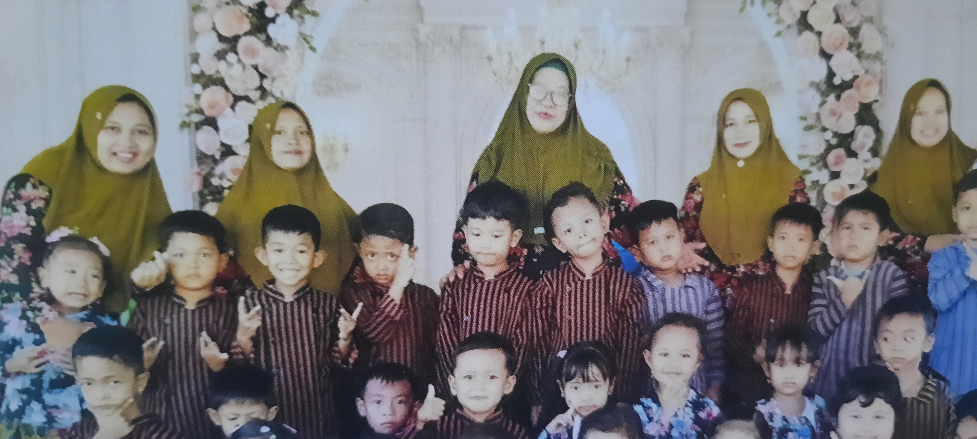 The image shows a group of people, primarily children and women, posing for a photograph in what appears to be an indoor setting with floral decorations. The background consists of a light-colored, possibly draped, fabric and large floral arrangements featuring pink and white flowers.
In the background, three women are visible, all wearing olive-green hijabs. The woman in the center is wearing glasses. The two women on the sides are also wearing patterned dresses or tops underneath their hijabs.
In the foreground and mid-ground, there are numerous children, mostly boys, dressed in matching dark-colored, striped outfits. Several children are smiling, while others have neutral or serious expressions. Some children are making hand gestures.
One girl in the front row on the left is wearing a floral patterned dress and has a pink hair accessory. Another girl in the center of the front row is wearing a similar floral dress.
The overall impression is that of a group photograph taken at a special event, possibly a school or family gathering, in Ploso, Indonesia.