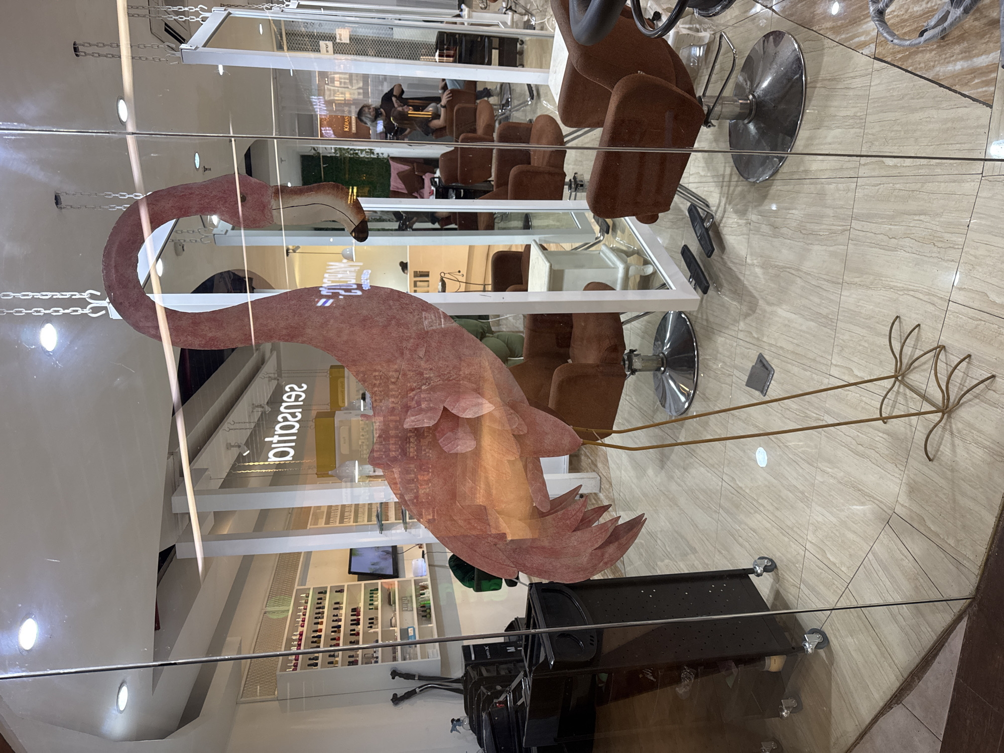 This image captures the interior of a hair salon or beauty parlor, viewed through a glass partition or window, resulting in prominent reflections. The scene is presented sideways and depicts an indoor setting with artificial lighting.
A large, decorative pink flamingo, seemingly crafted from layered paper or fabric, dominates the foreground. Its long, curved neck and beak point towards the left, while its body extends to the right, supported by thin, gold-colored rod legs. This flamingo adds a whimsical and distinctive element to the salon's facade or interior display.
Inside the salon, several brown, upholstered salon chairs with chrome bases are visible, arranged in rows. A black, mobile salon trolley with wheels is positioned in the lower right foreground. The floor is tiled with a light, patterned material. In the background, white shelving units are stocked with numerous bottles and beauty products, possibly hair care items or nail polishes. A television screen is also visible on one of the shelves.
Activity within the salon includes at least one person, likely a stylist, attending to a client seated in one of the chairs. Other reflections on the glass further suggest the presence of people, but their details are obscured.
Notable details include the heavy reflections on the glass, which superimpose parts of the exterior environment onto the salon interior. The word "Sensation" is visible on a wall inside the salon, appearing reversed due to the reflection. The overall impression is that of a modern and well-appointed beauty establishment. The location context provided is Kuta Selatan, Indonesia.