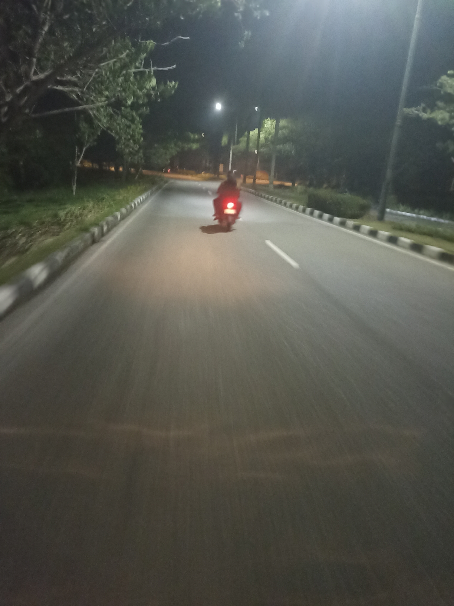A lone motorcyclist rides down a paved road at night. The motorcycle's tail light is illuminated red, and its headlight casts a beam ahead, visible against the dark asphalt. The road is lined with curbs that have a black and white striped pattern. To the left of the road, there is a grassy area with trees and foliage. To the right, more trees and what appear to be streetlights are visible. The overall scene is dark, with artificial lighting from the streetlights and the motorcycle creating illuminated areas on the road. This is a scene in Sungai Mempura, Indonesia.