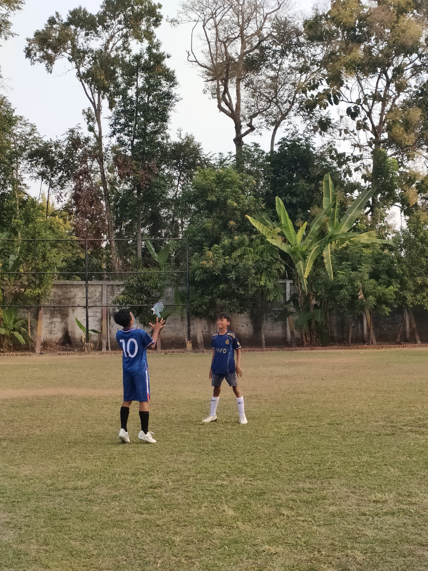 Two young boys in soccer uniforms are on a grassy field, likely part of the Banguntapan city area in Indonesia, given the visual cues of tropical trees and vegetation. One boy, wearing a blue uniform with the number 10 on the back, is in the foreground, tossing a white object, possibly a bottle or a ball, upwards with his right hand. He is facing towards the other boy, who is wearing a dark blue soccer uniform with "AFD" visible on the chest. This second boy is standing to the right and slightly behind the first boy, looking towards the object in the air. The field appears to be a sports ground, with a low concrete wall and a black metal fence in the background, backed by lush trees and foliage. The sky is overcast.