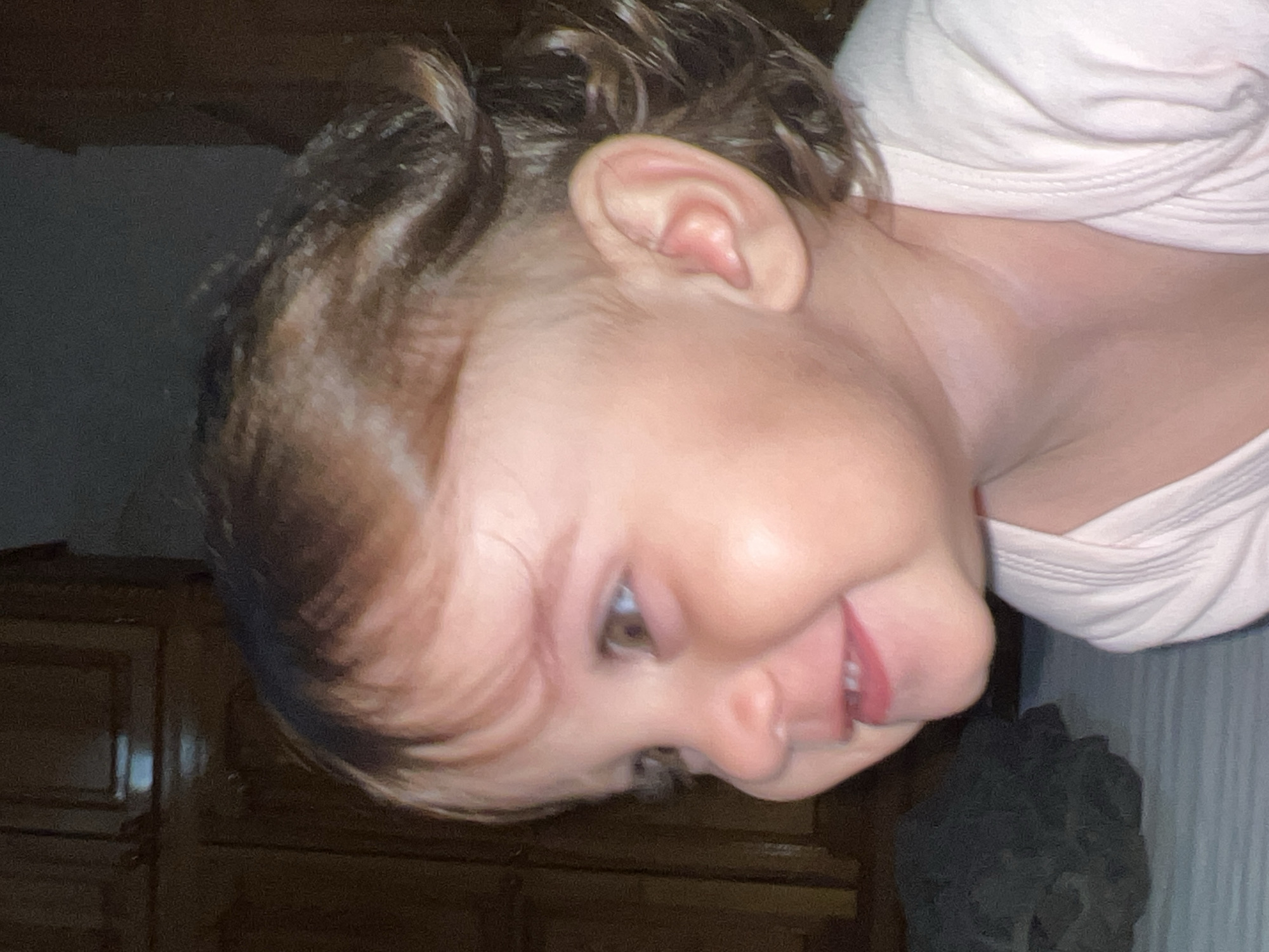 A close-up, slightly blurred image shows a baby or young child lying down and turned to their side, facing the camera. The child has fair skin, brown hair with some curls, and visible ear. They are wearing a white or light pink top. The background is dark and somewhat indistinct, but includes a wooden cabinet or dresser. Location in Elena, Bulgaria is not supported by the given images.
