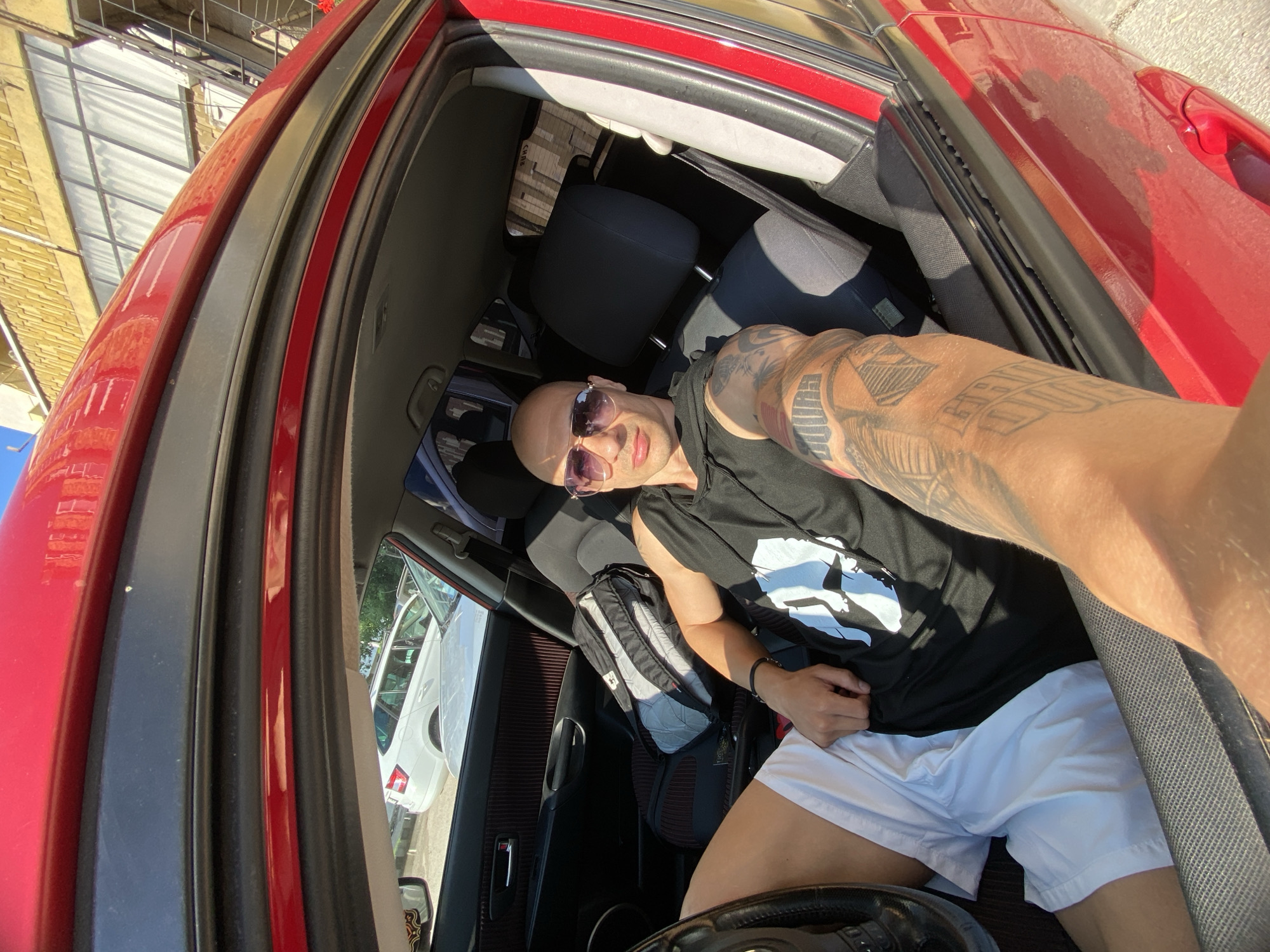 The image is a selfie taken by a bald man with extensively tattooed arms, seated in the driver's seat of a red car. He is wearing dark sunglasses, a black t-shirt with a white graphic, and white shorts, and has a neutral to slightly smiling expression while looking directly at the camera. The image is oriented sideways, with the man's head facing the left of the frame.
The setting is the interior of a car, specifically the front cabin, illuminated by bright daylight, suggesting a sunny day with clear skies. Strong shadows are cast within the car. Through the windows, parts of an urban environment are visible, including a building with light-colored siding and another white car parked nearby.
A black and grey backpack is placed on the front passenger seat. A small portion of the steering wheel is visible in the lower part of the frame. The tattoos on the man's arms feature intricate designs, including stylized letters and geometric patterns, though specific words are not clearly legible. A faint label, possibly reading "CARE," is visible on the headrest of the passenger seat. The overall scene suggests a casual moment captured in a car, possibly while parked, with no overt activity taking place other than the selfie itself. No explicit visual cues within the image confirm the location as Pleven, Bulgaria.