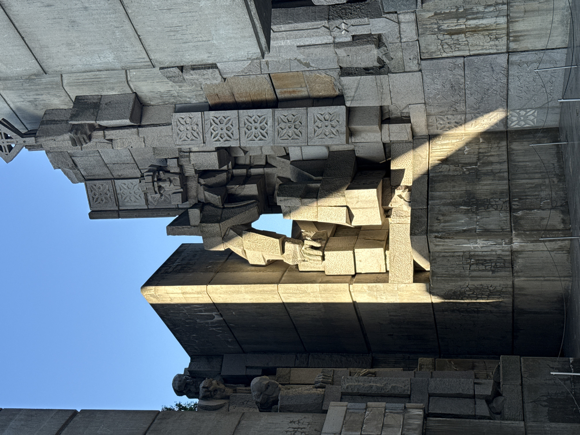 The image shows an upward view of a section of the Monument to 1300 Years of Bulgaria, located in Shumen, Bulgaria. The monument's architecture is characterized by large, geometric concrete blocks, some adorned with repeating rosette patterns. A beam of sunlight illuminates a portion of the structure, creating a contrast between light and shadow. The sky is visible in the gaps between the blocks, appearing as a clear, pale blue. The architectural details include stylized heads or gargoyle-like features along a lower edge of the monument. The photograph is taken from a low angle, emphasizing the monument's scale and complexity.