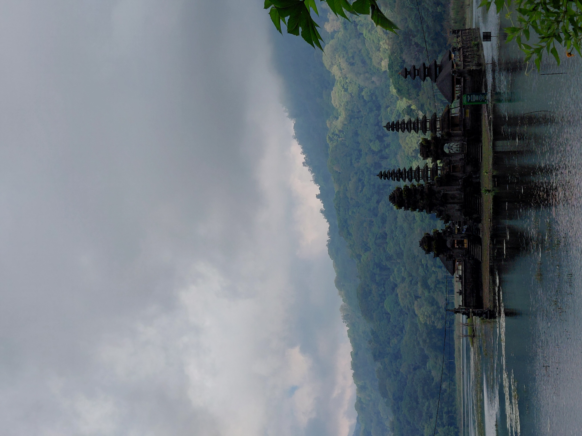 A body of water, likely a lake, is in the foreground and middle ground of the image, with some vegetation floating on its surface. In the center of the image, an island structure with several tiered pagodas, a traditional Balinese temple complex, is situated in the water. The temple is dark stone and appears ancient. Behind the temple, lush green, mist-covered hills rise, indicating a mountainous or hilly terrain. The sky above is overcast with thick, gray clouds. The scene is calm and serene, with the temple's reflection visible in the water. The location is Pura Ulun Danu Bratan, a famous Hindu temple situated on the shores of Lake Bratan in the mountains near Bedugul, Bali, Indonesia. The image captures the temple and its surrounding natural beauty under a cloudy sky.