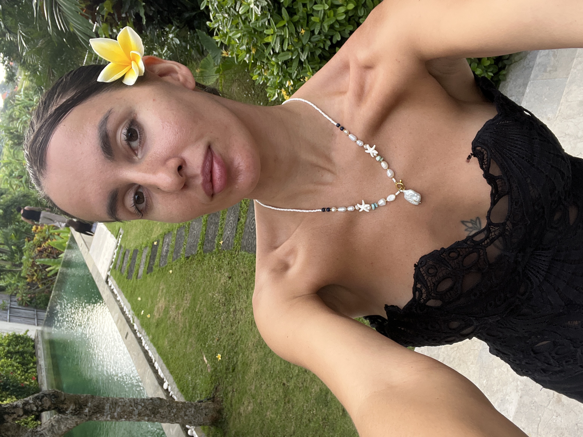 A woman is posing for a selfie in what appears to be a tropical setting in North Kuta, Indonesia. She is wearing a black lace top and a necklace with pearls and starfish charms. She also has a yellow flower in her hair. Behind her, there is a lawn, a pool, and lush greenery.