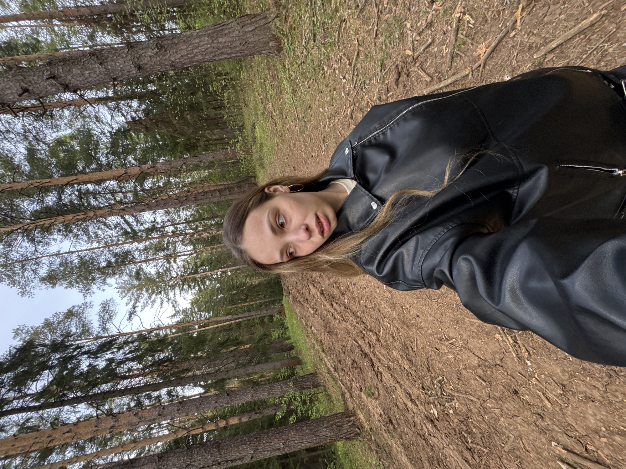 A woman with long blonde hair wearing a black leather jacket is pictured in a wooded area in Прикамский, Russia. Tall pine trees surround her, their trunks visible on the left side of the frame. The ground is covered in brown dirt and scattered sticks and foliage. The sky is visible through the trees, appearing as a light gray. The image has been rotated counter-clockwise.