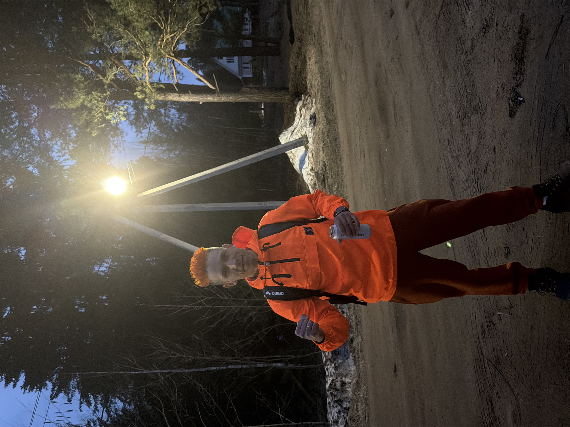 A man stands on a dirt road near what appears to be a snowy patch and some trees. He is illuminated by a nearby streetlamp. The man is wearing an orange jacket, reddish-brown pants, and dark shoes. He holds a can in his right hand and has a backpack on. The environment suggests a rural or suburban area in Chaikovsky, Russia, with sparse vegetation and a house visible in the background.